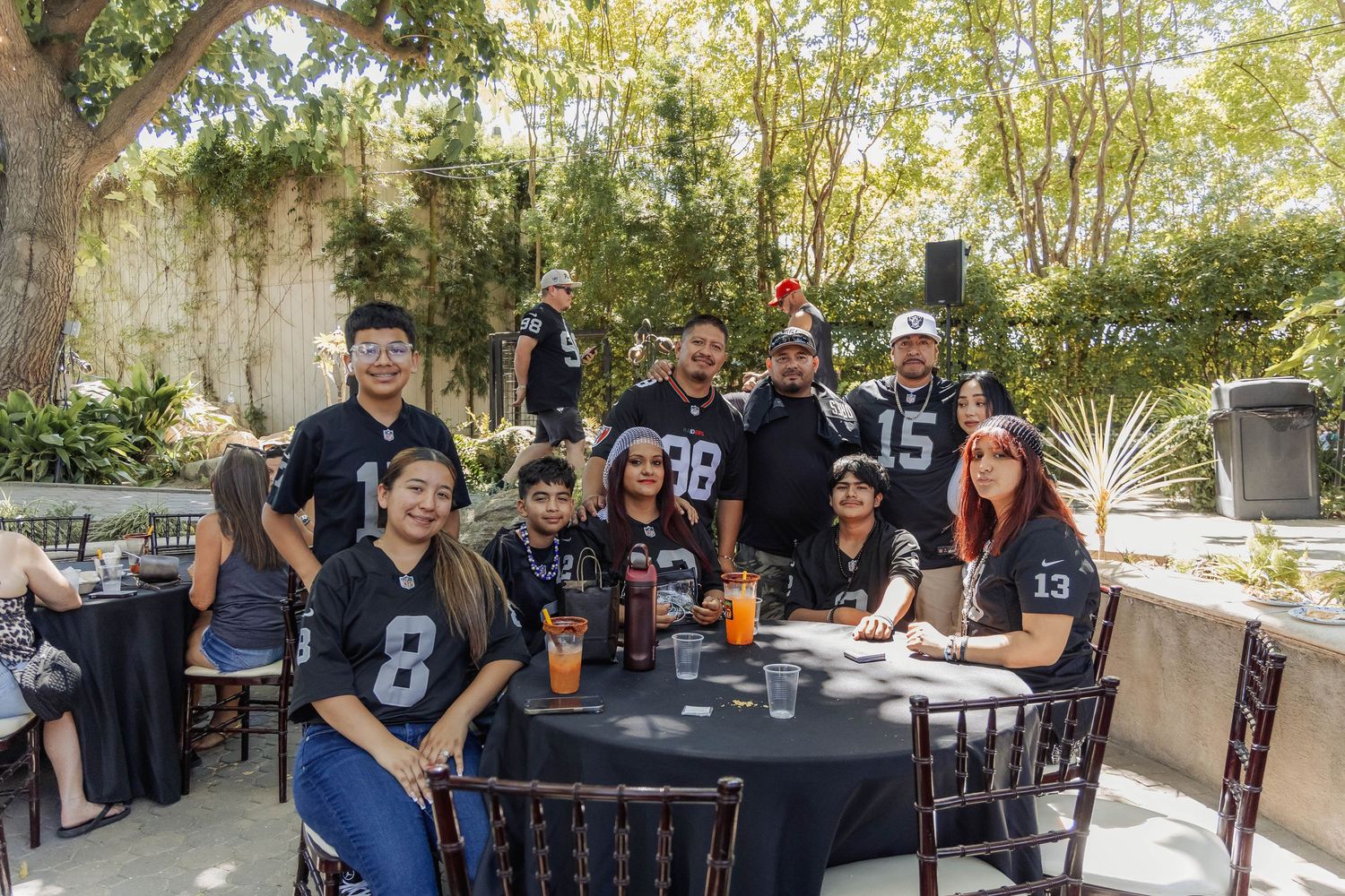 Group of Raiders NFL fans in black jerseys gathered around table at outdoor fan event.