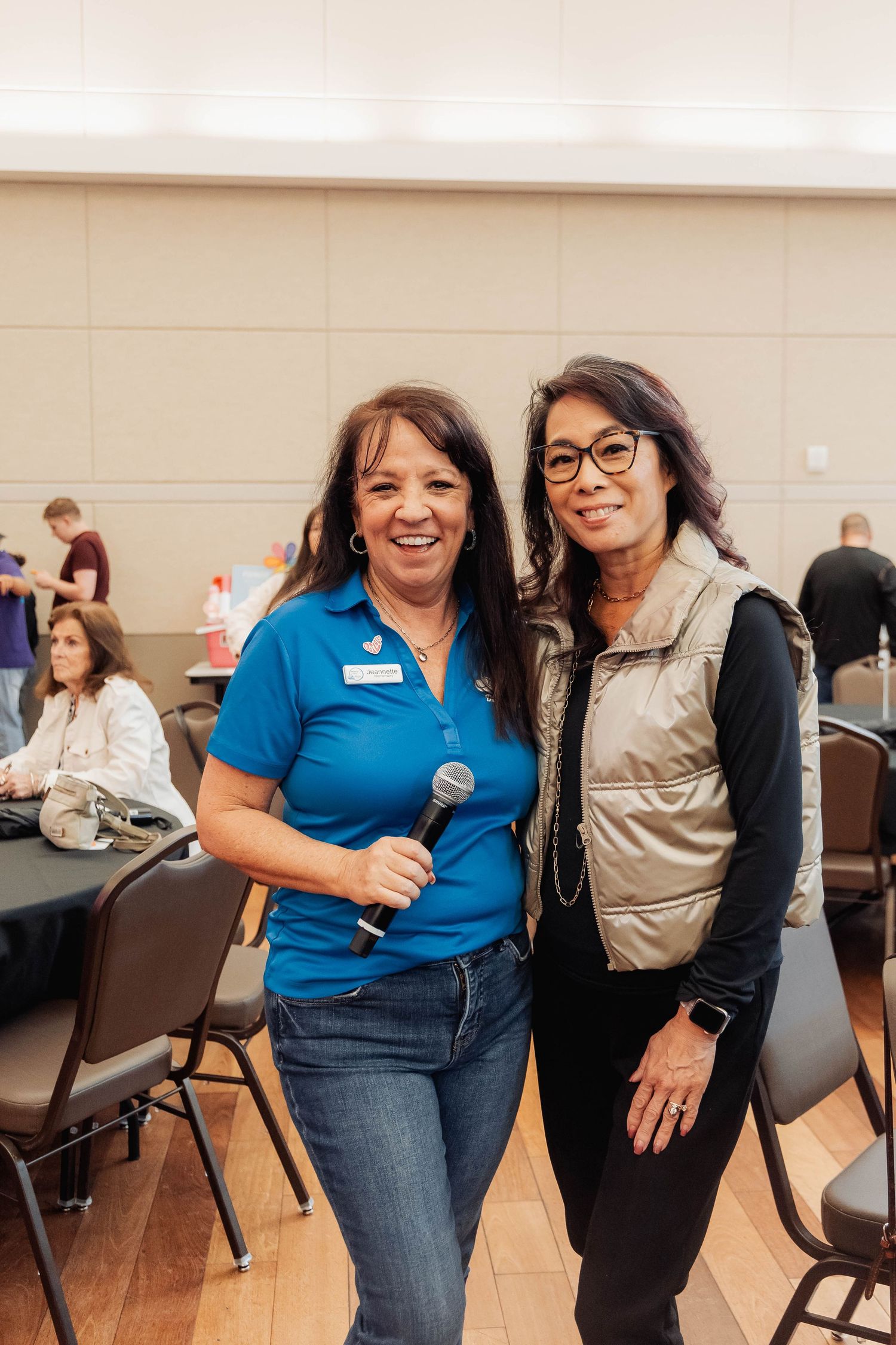 Two women smiling together at an indoor event, one holding a microphone and wearing a blue polo shirt.