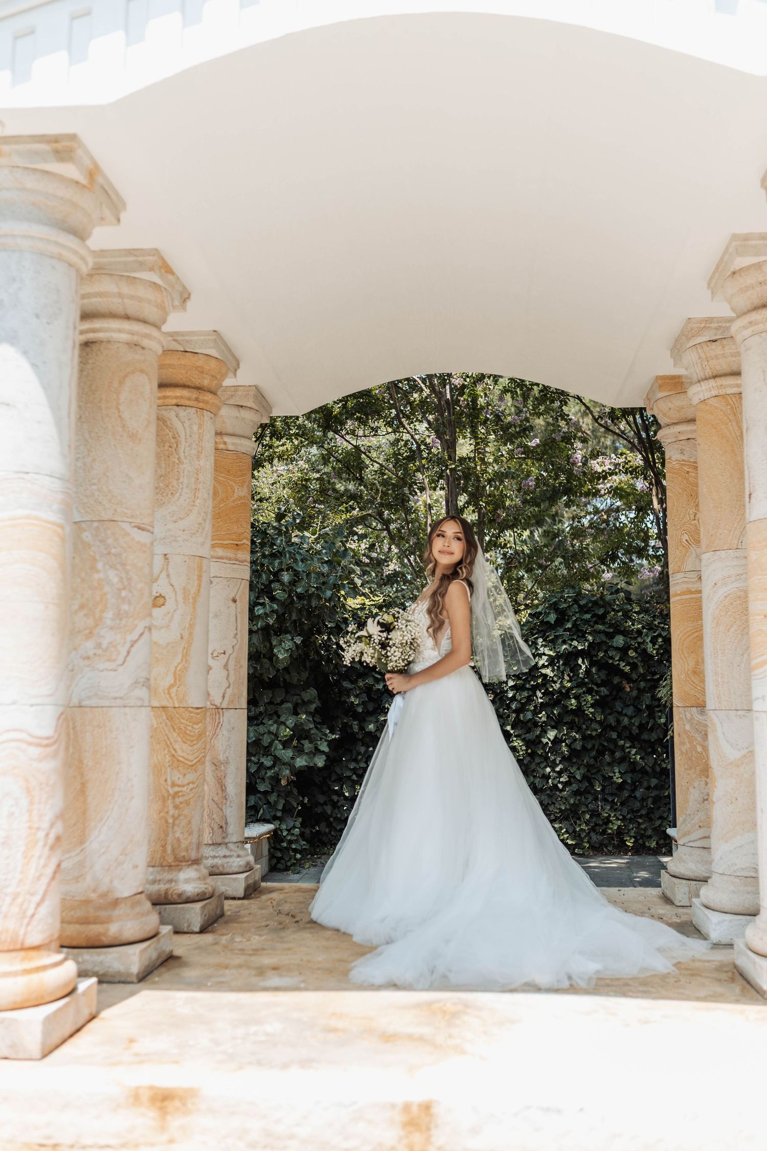 A bride in a flowing white ballgown poses under stone archways in a romantic garden setting.