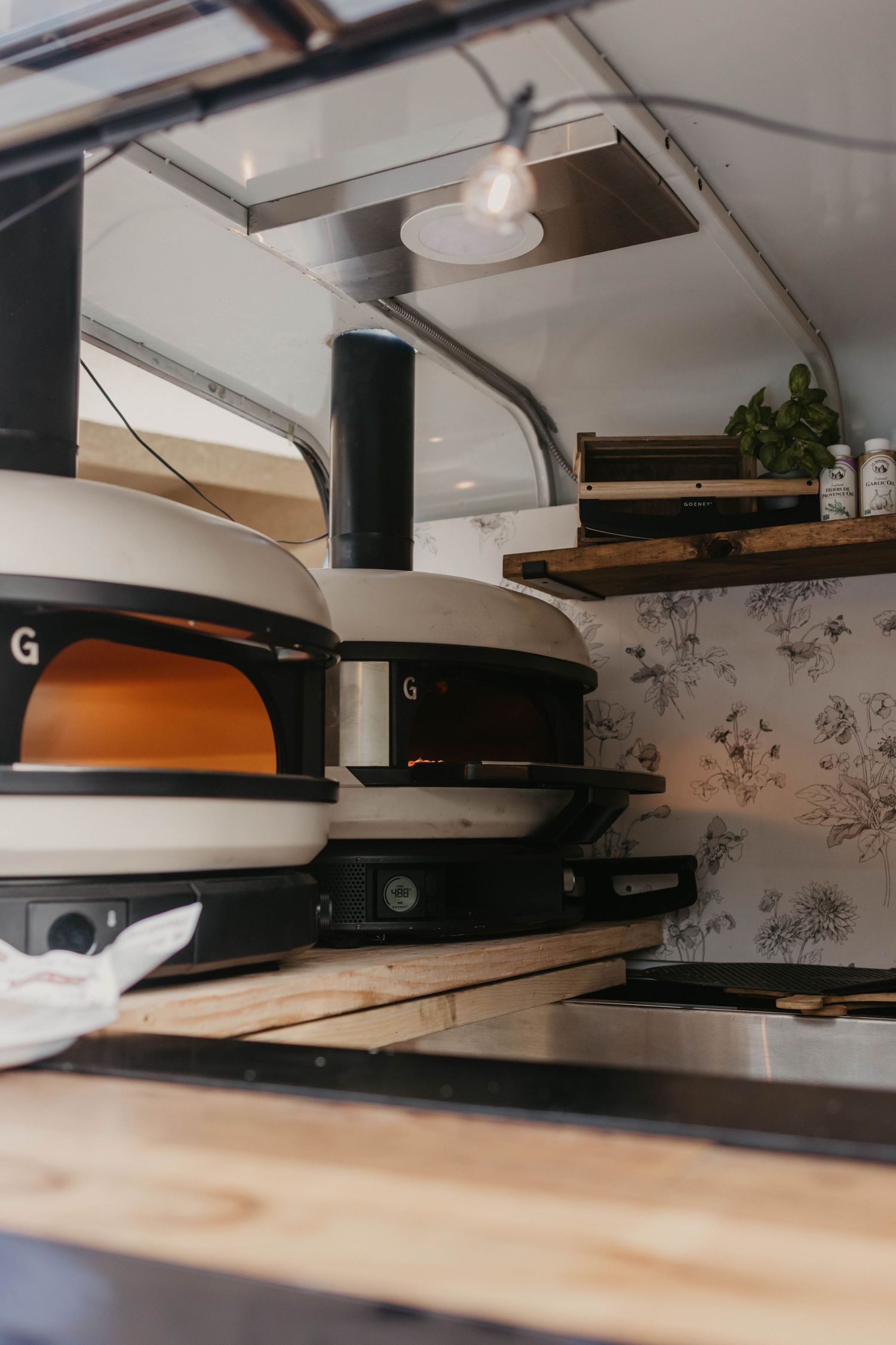 Commercial pizza ovens mounted in a kitchen with white walls and floral wallpaper.
