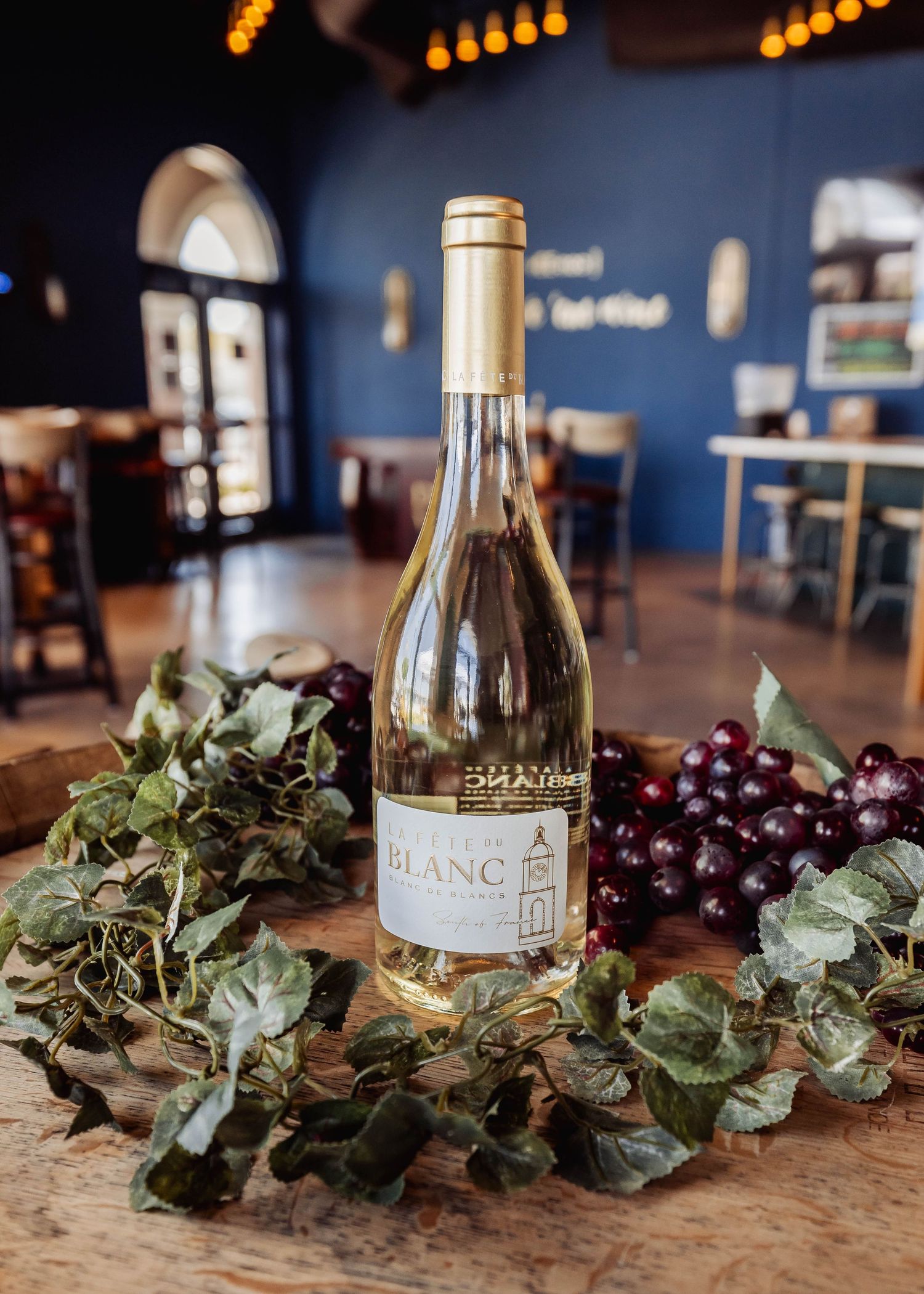 A bottle of white wine sits on a rustic wooden table with decorative greenery and fresh purple grapes.