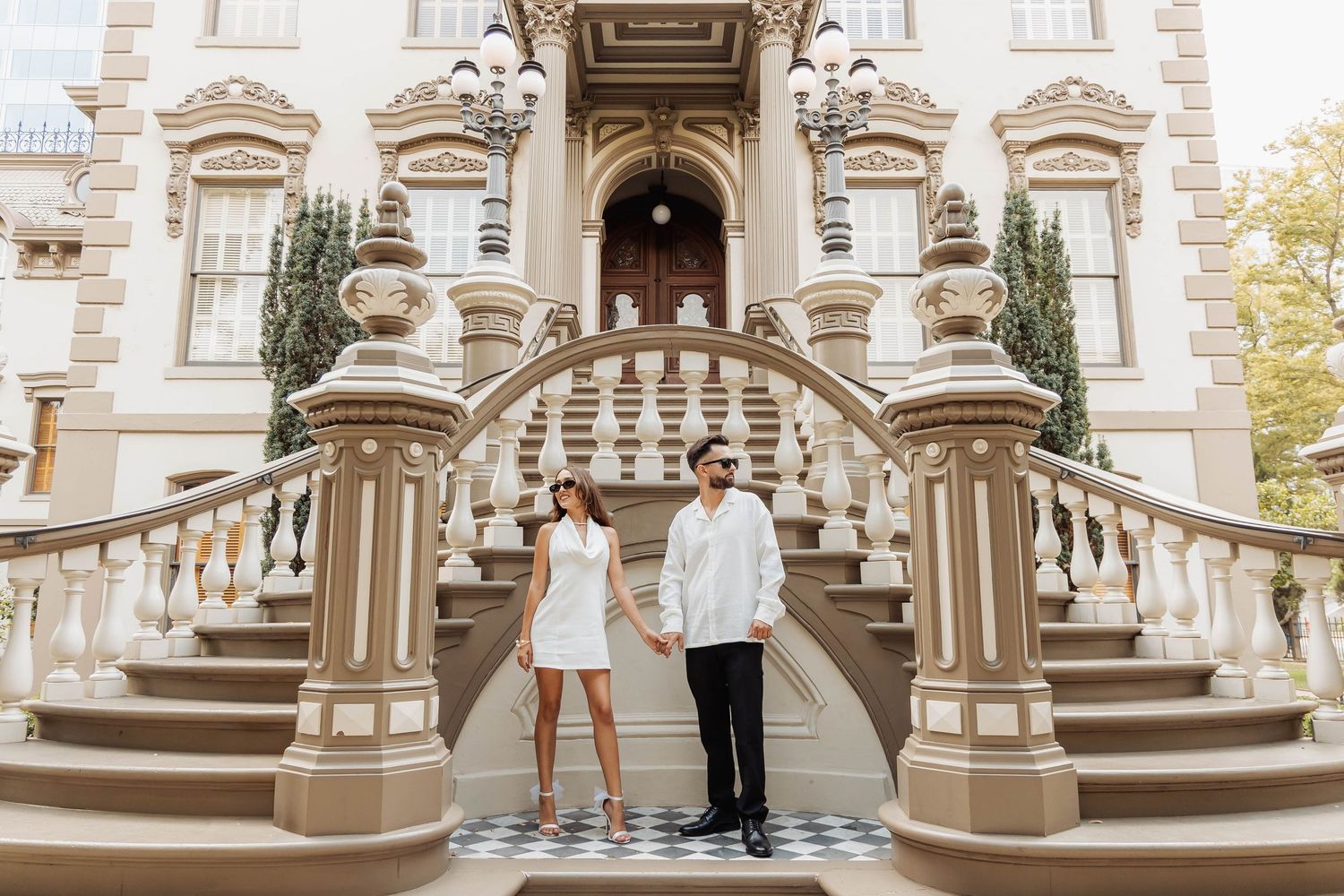 Couple in matching white outfits standing on grand mansion steps with ornate architecture.