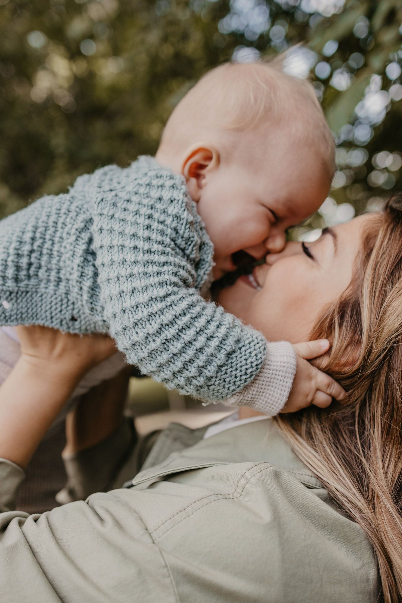 Intimate close-up moment between parent and baby wearing knit sweater against natural outdoor backdrop.