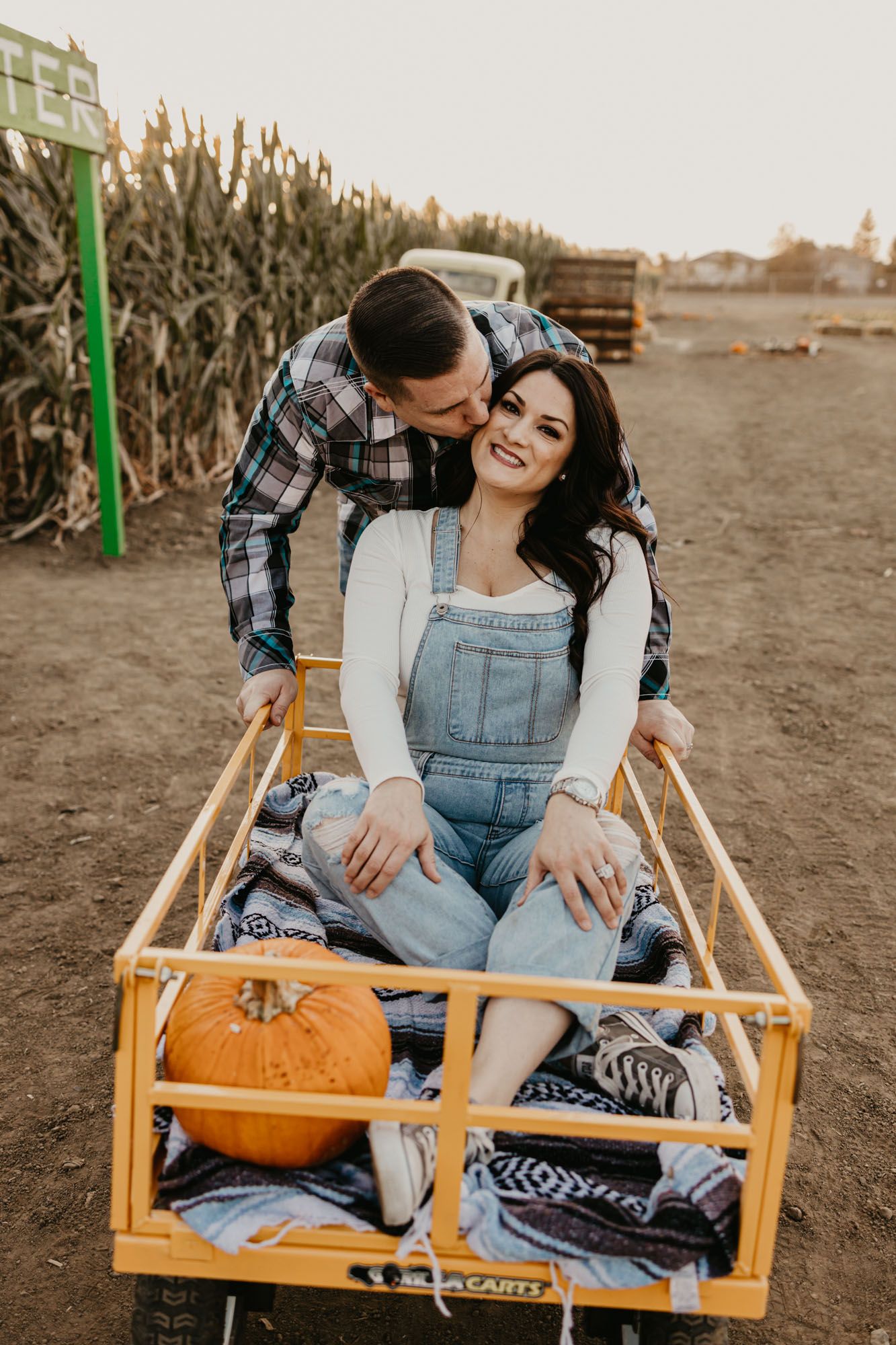 A couple sharing a tender moment in a rustic wagon beside autumn corn stalks.