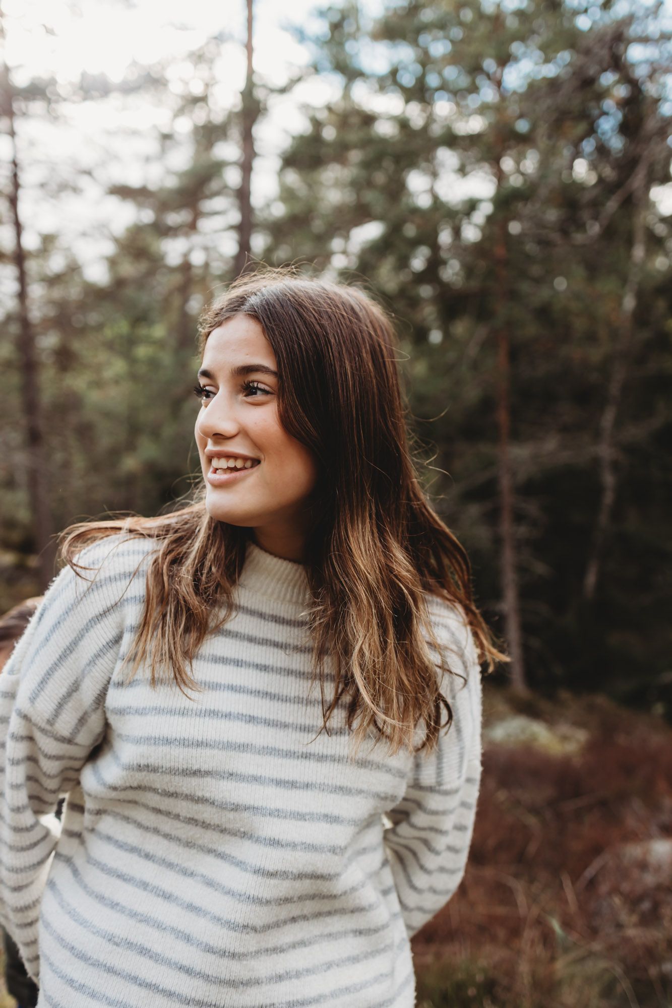 Person in striped sweater smiling while standing in front of pine forest.