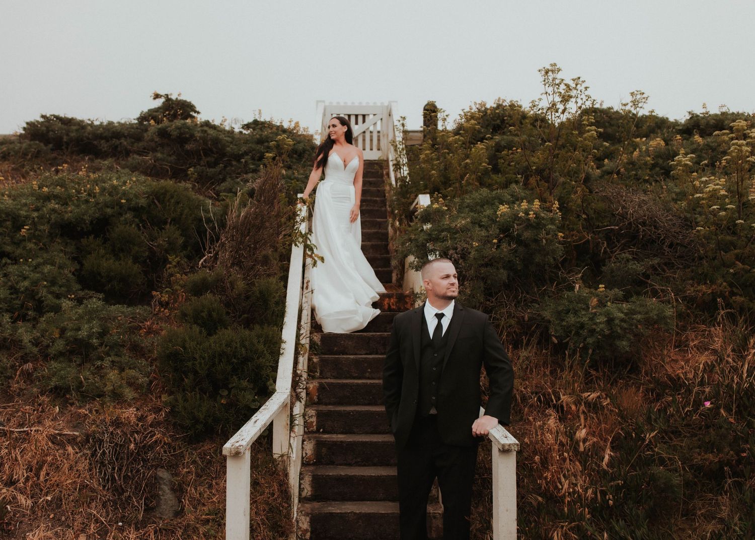 A moody wedding photo showing a couple on rustic wooden stairs along a hillside path.
