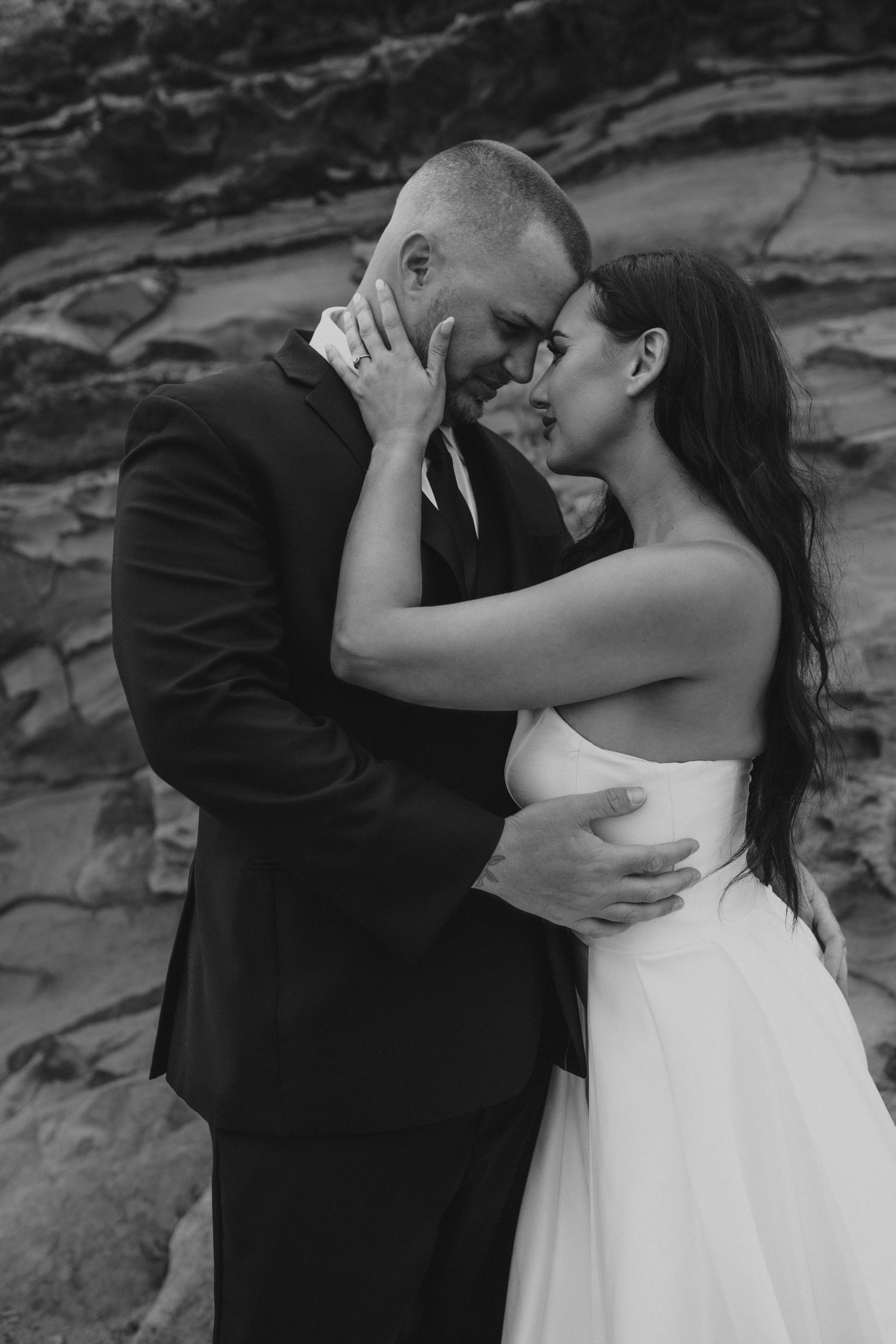 Black and white intimate portrait of newlyweds embracing against textured rock wall.