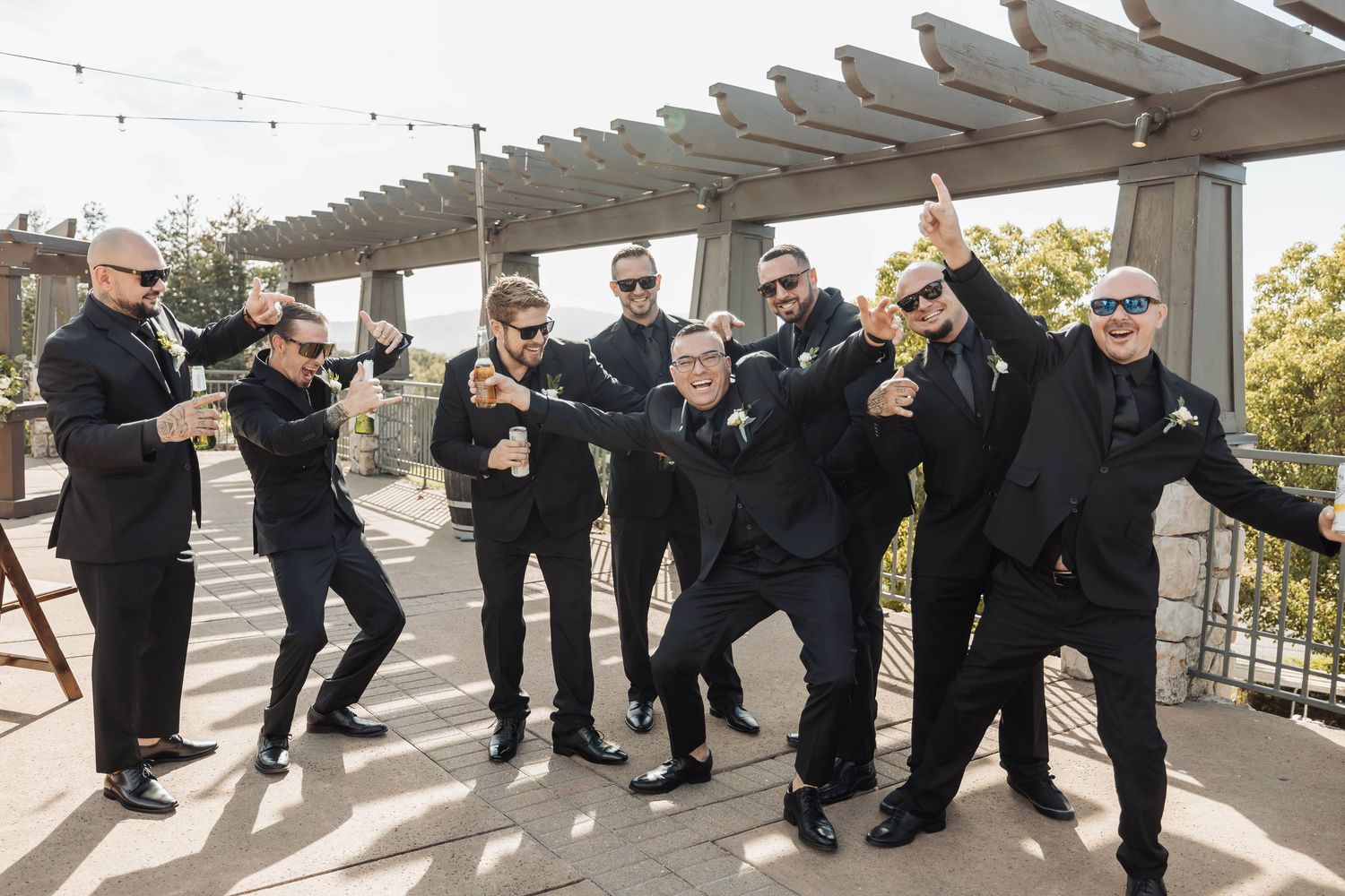 Groomsmen in black suits strike fun poses under outdoor pergola.