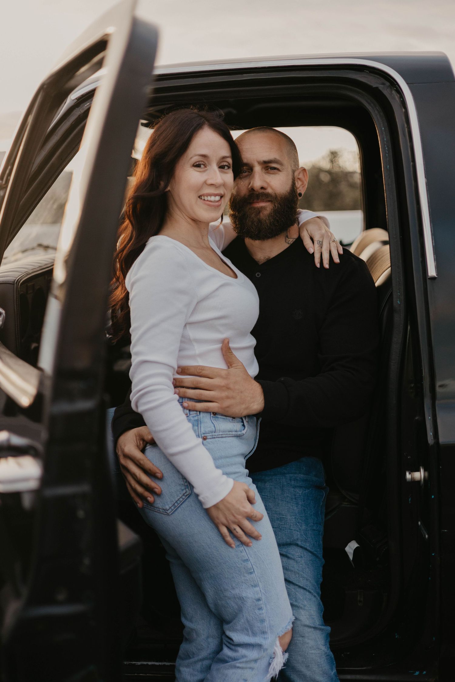 Affectionate couple embracing by car door in casual denim and white outfits.