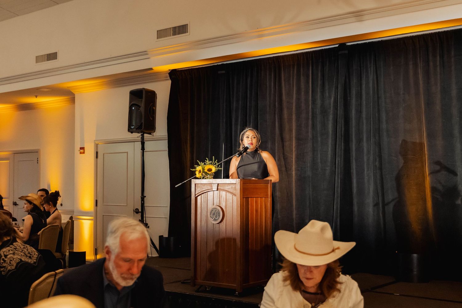 Woman speaking at podium on stage at formal event with audience in foreground and dark curtain backdrop.