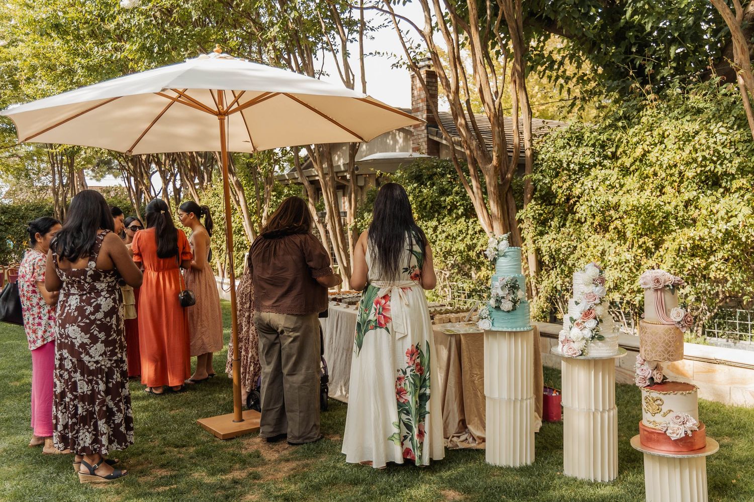 Outdoor wedding cake display with tiered cakes on pedestals, guests mingling under a patio umbrella in a lush garden.