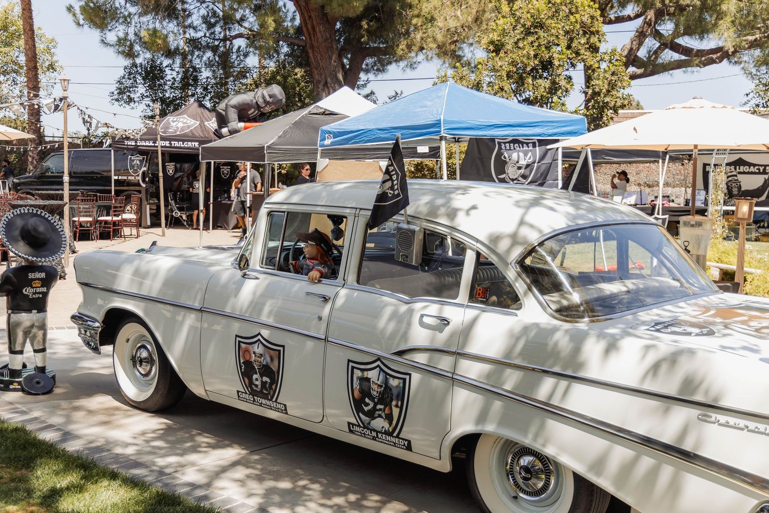 Vintage white 1957 Chevy car with Raiders logo at an outdoor tailgate party under blue canopy tents.