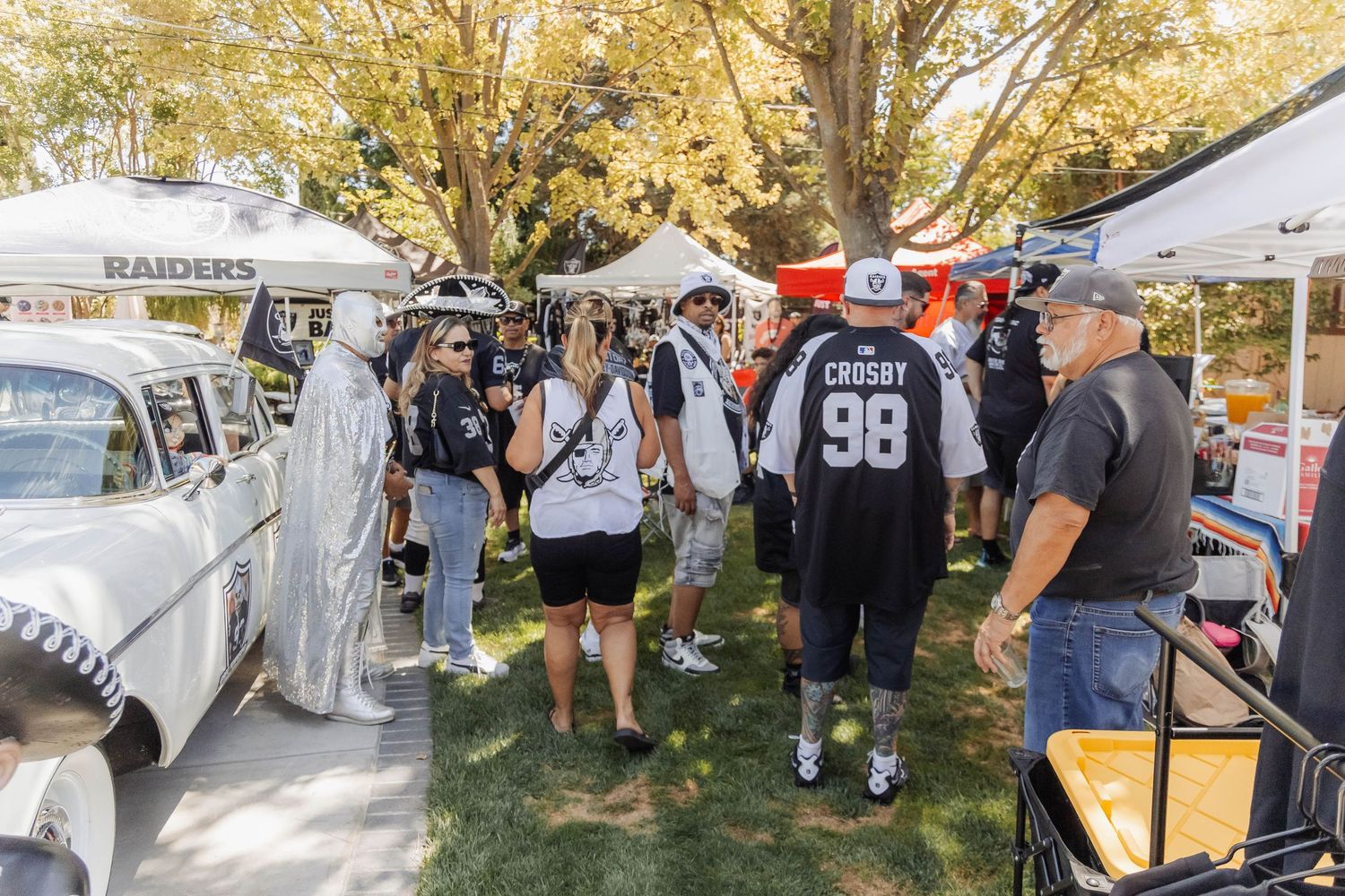 Raiders fans in black and white jerseys mingling among vendor tents and classic cars at an outdoor tailgate event.