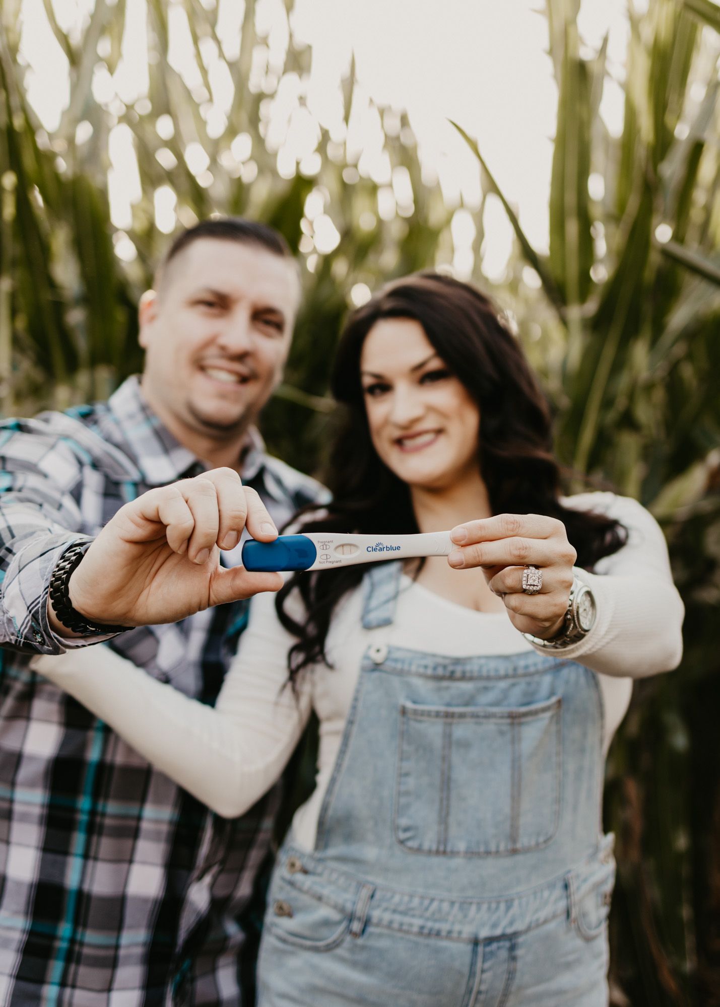 Happy couple displaying positive pregnancy test against cornfield background during announcement shoot.