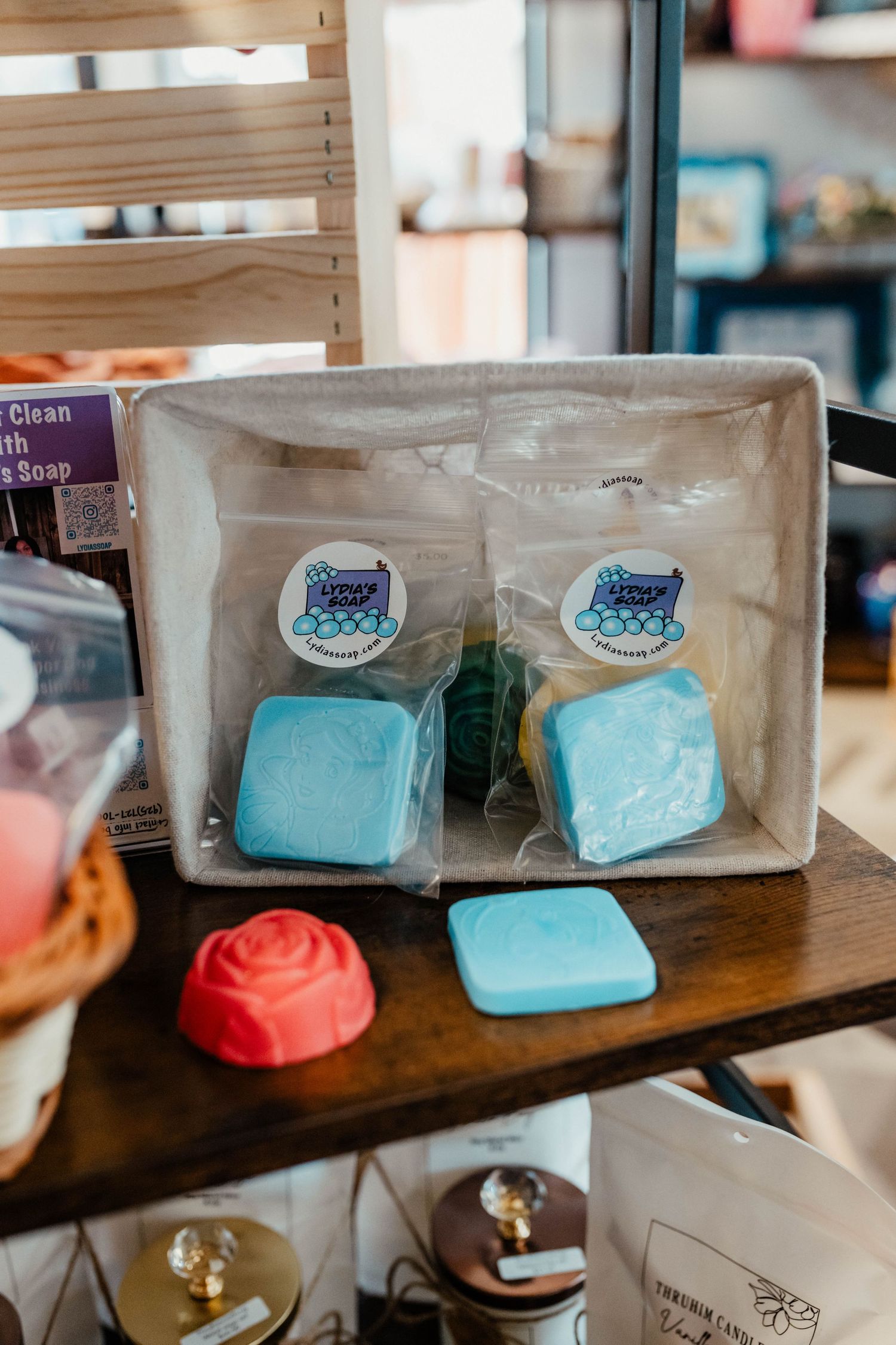 Blue square soaps with cute cat logo stickers displayed in clear packaging next to a pink rose-shaped item on wooden surface.