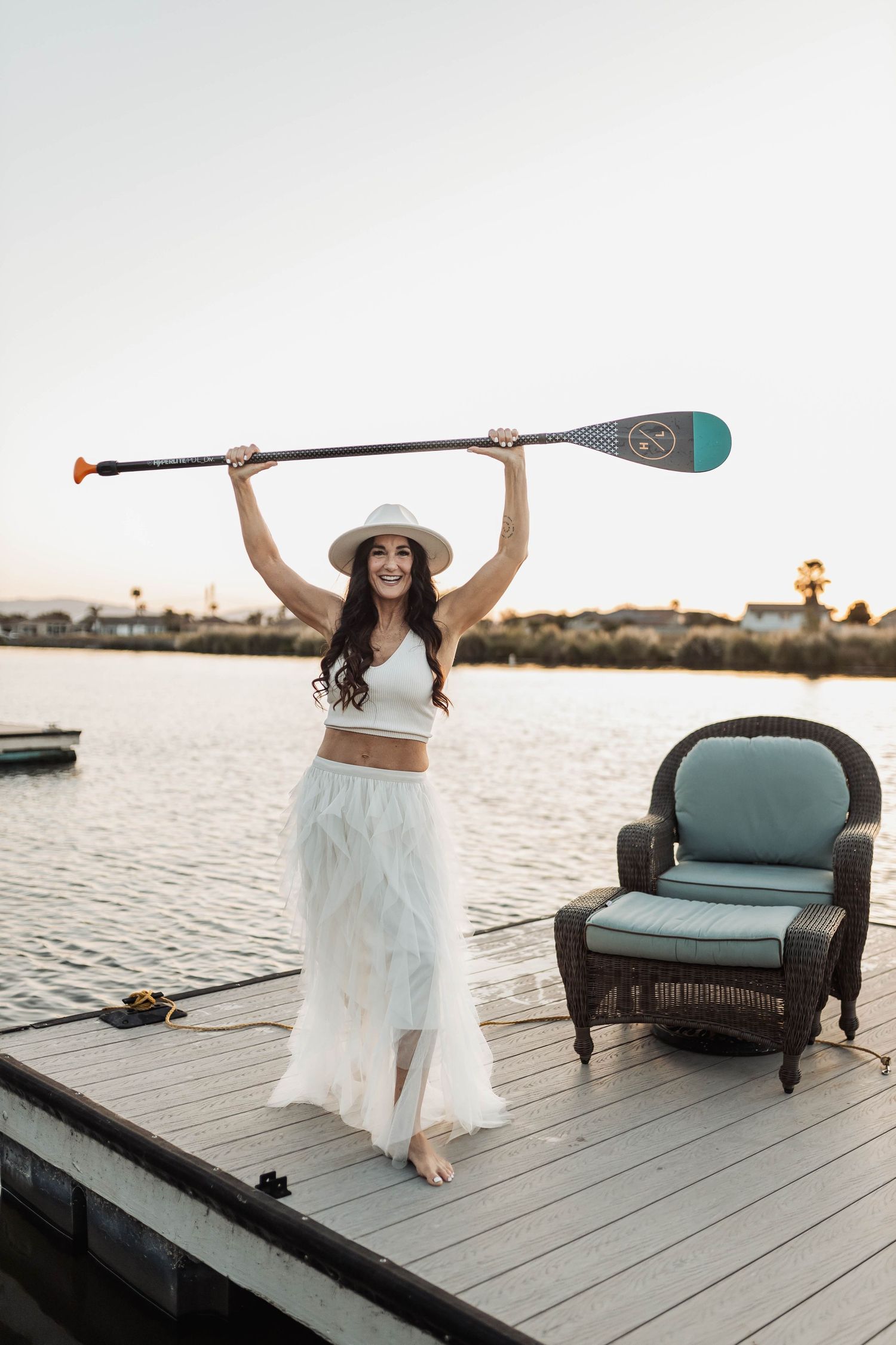 Woman in white dress holding paddle overhead on dock with teal chair.