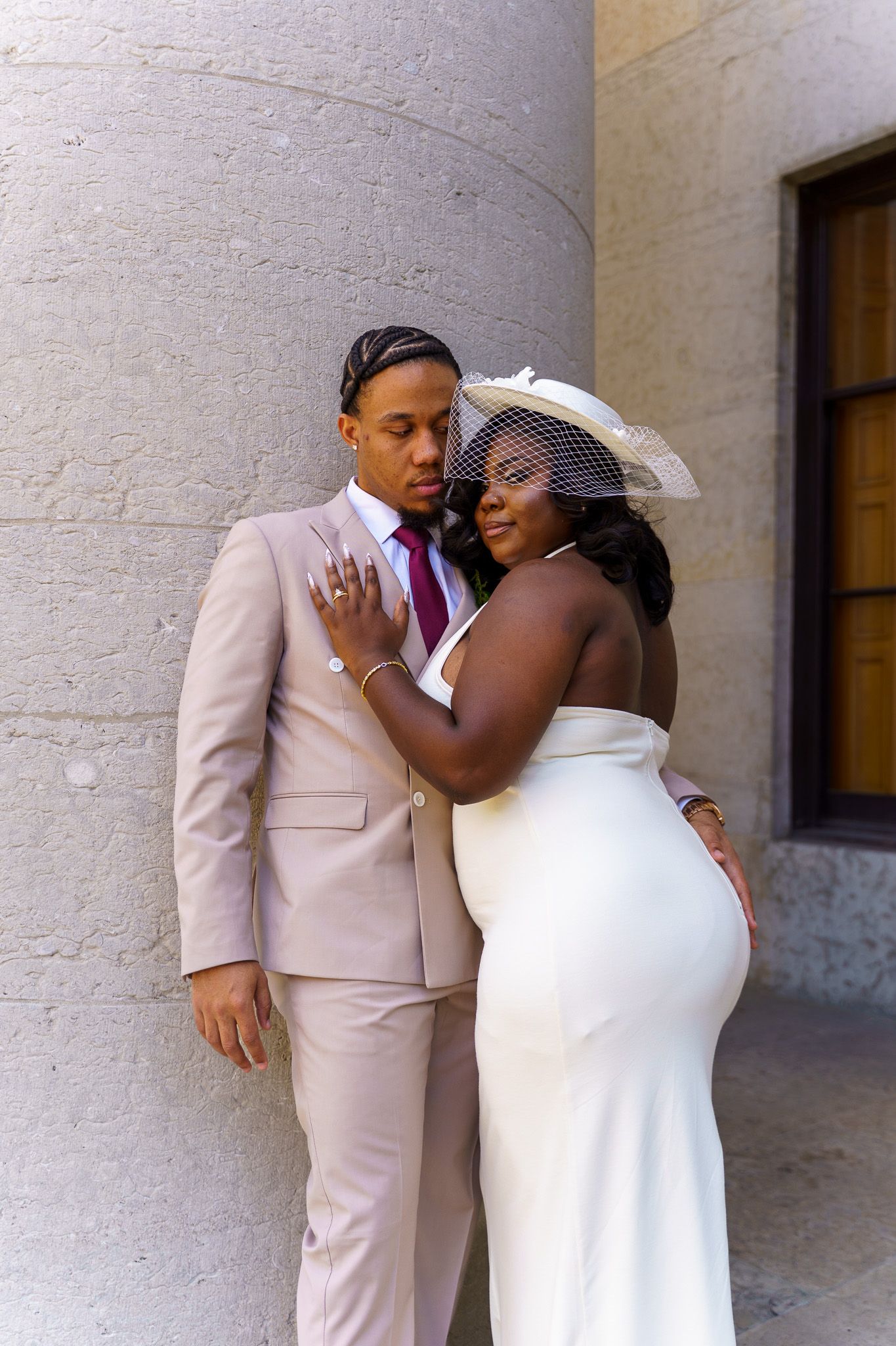 A couple in wedding attire poses by a stone column, groom in blush suit, bride in white gown and birdcage veil hat.