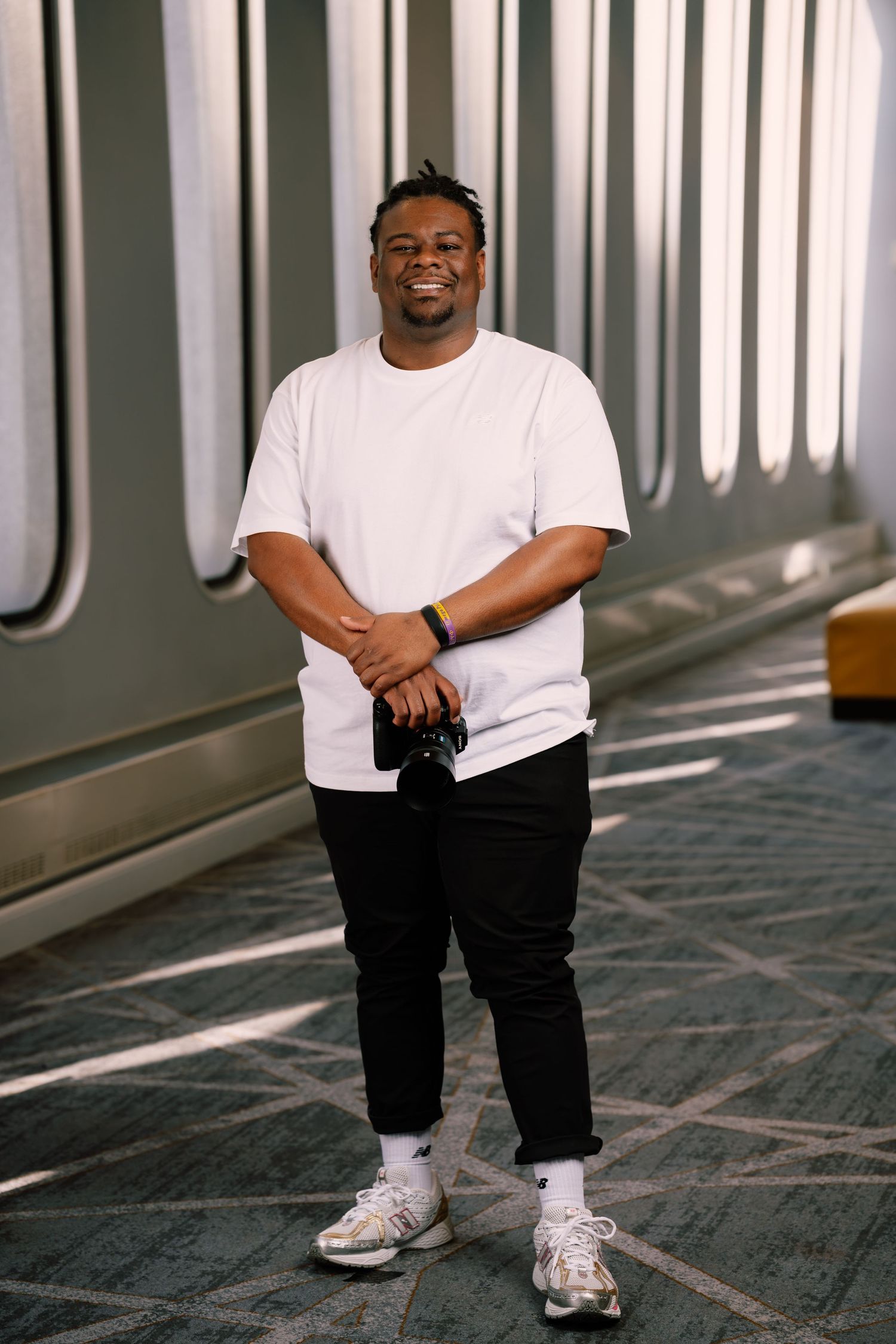 Smiling man in white t-shirt and black pants holds a camera while standing in a modern corridor.
