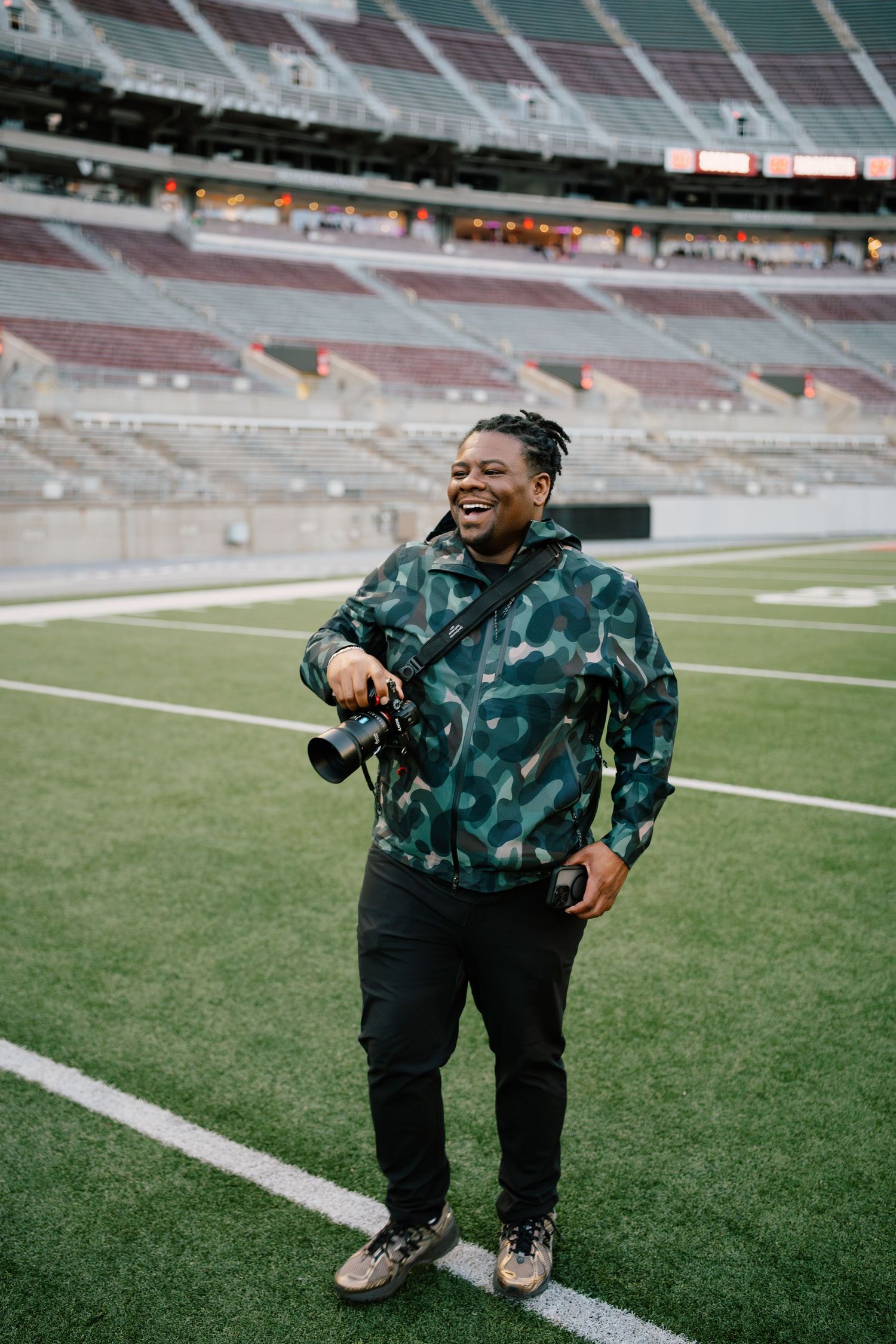 Smiling photographer holding camera on football field sideline wearing camo hoodie and black pants in empty stadium.