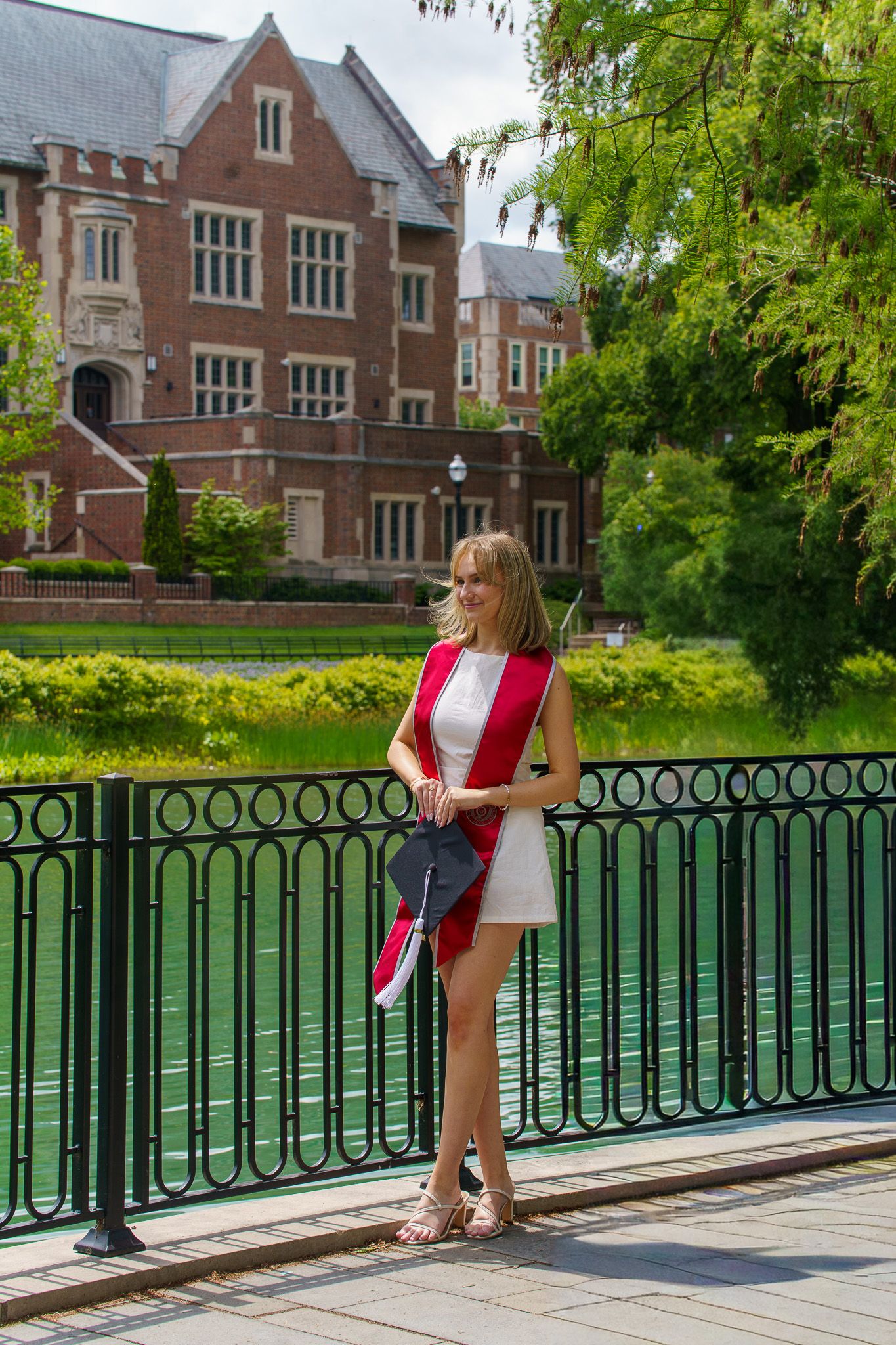 A young woman in a white dress and red graduation sash poses by a railing near a pond on a college campus.