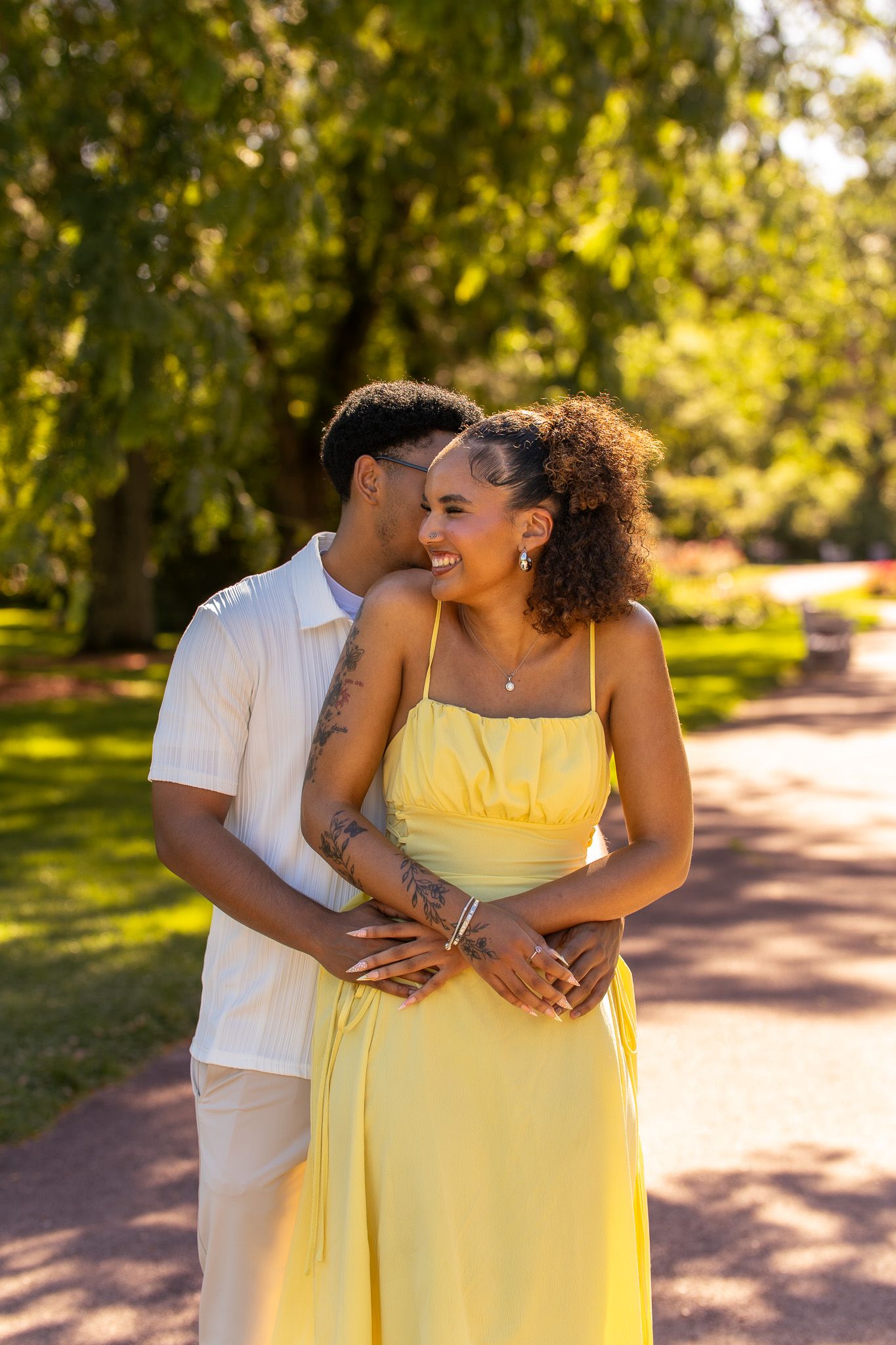 A couple embraces in a sunny park, woman in yellow dress smiling as man kisses her cheek.