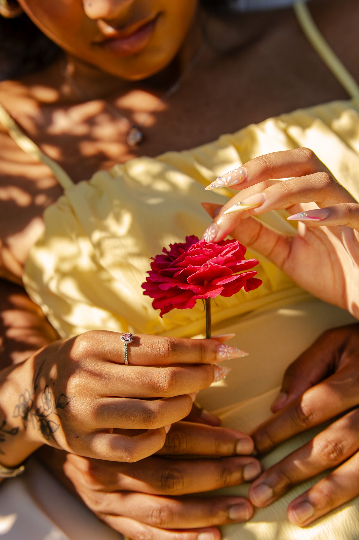 A woman with manicured nails and a diamond ring holds a red carnation flower, wearing a yellow dress in warm sunlight.