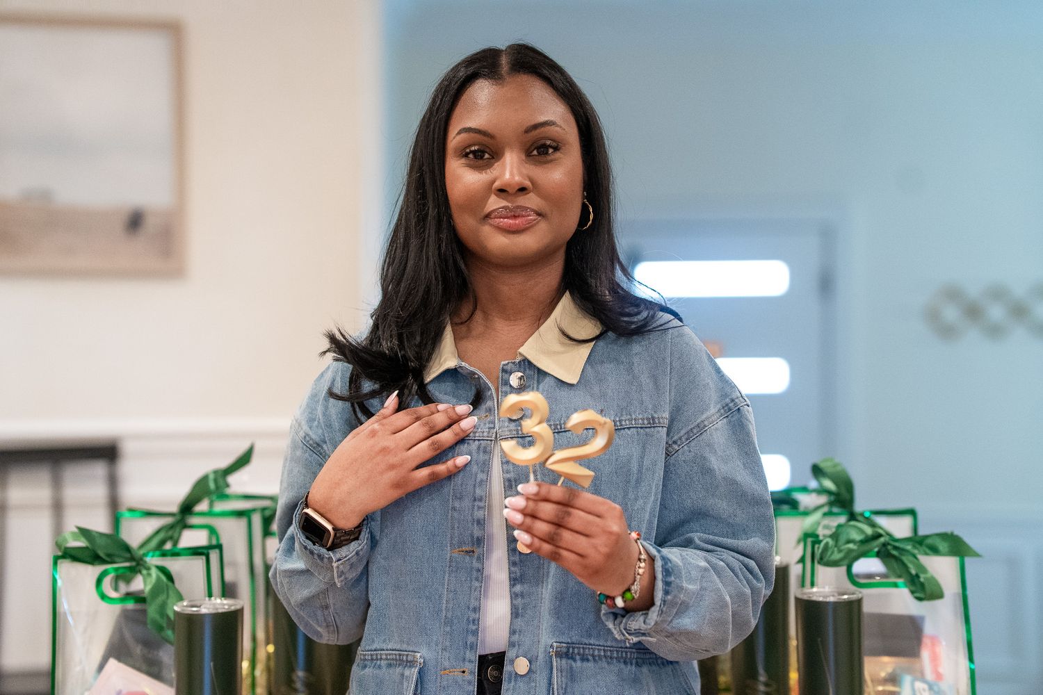 Woman in denim jacket holding a sparkly number 32 candle, smiling at a birthday celebration indoors.