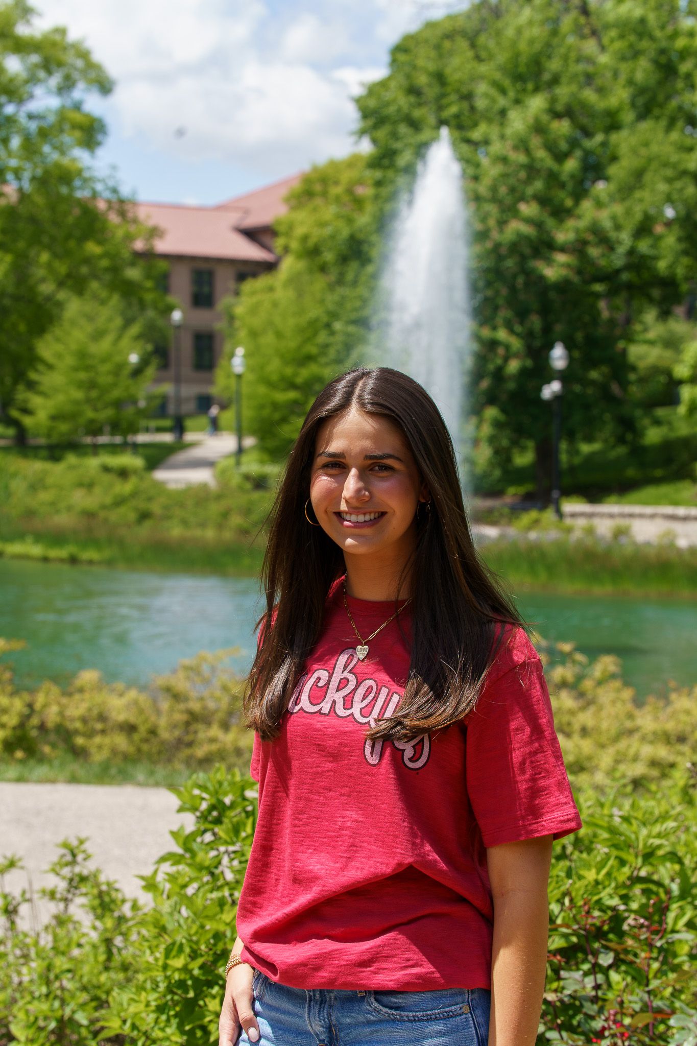 Young woman in red Buckeyes t-shirt smiling outdoors near a fountain and pond on a sunny day.