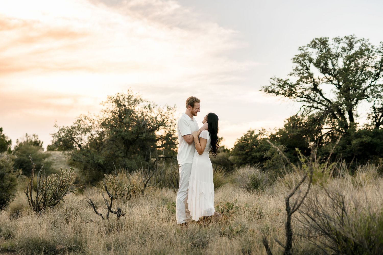 Couple in white attire embracing at sunset in a desert field with golden grasses and trees.