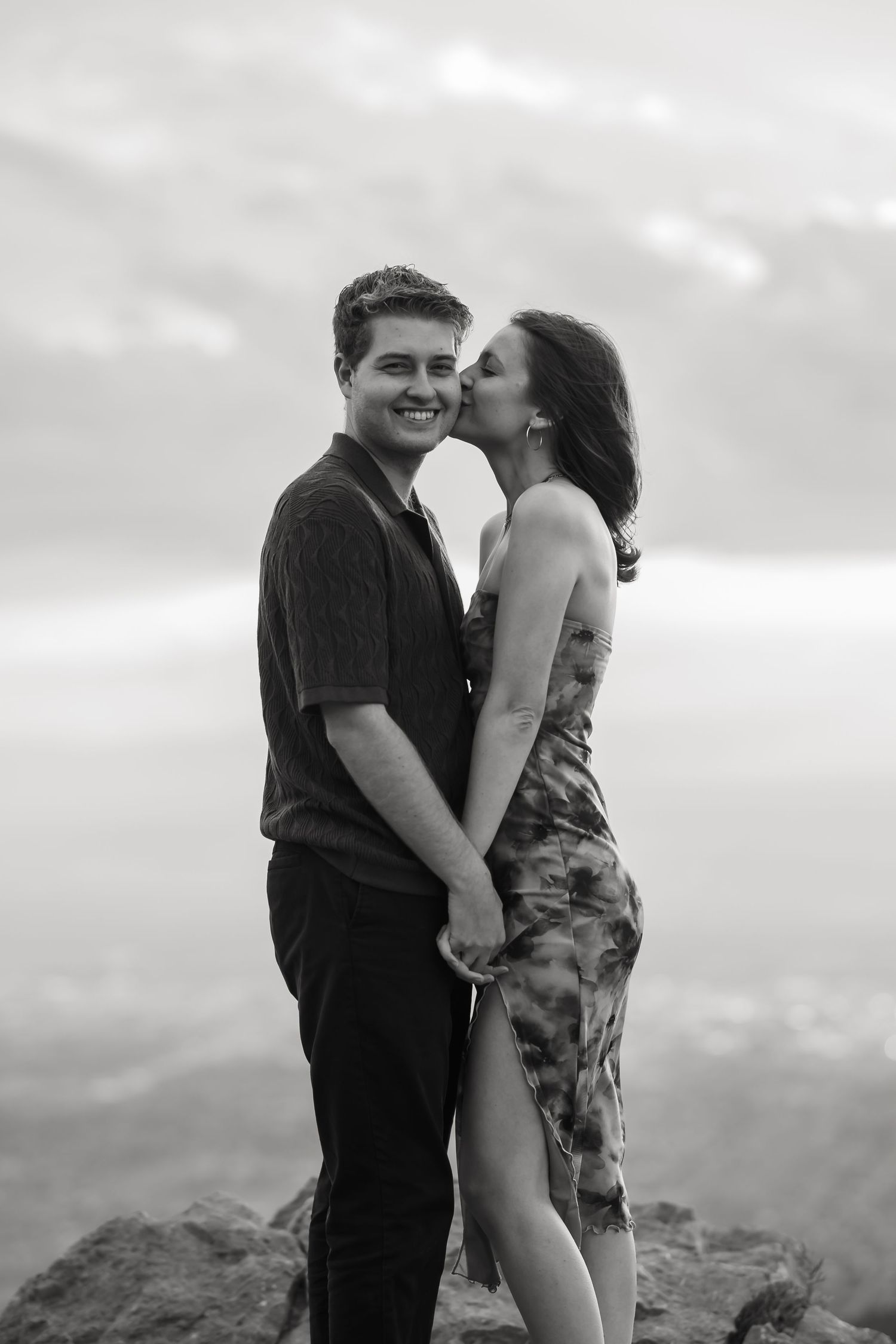 A couple embraces while sitting on a rocky cliff edge with a scenic vista in the background.