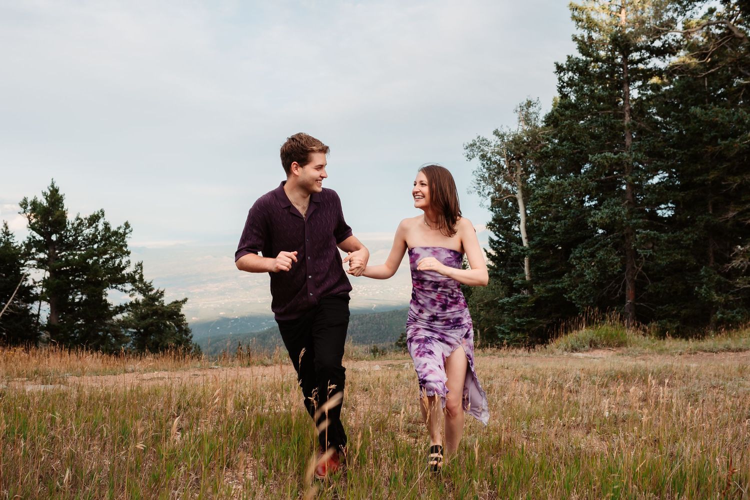 Couple running together through a mountain meadow surrounded by evergreen trees and rolling hills.