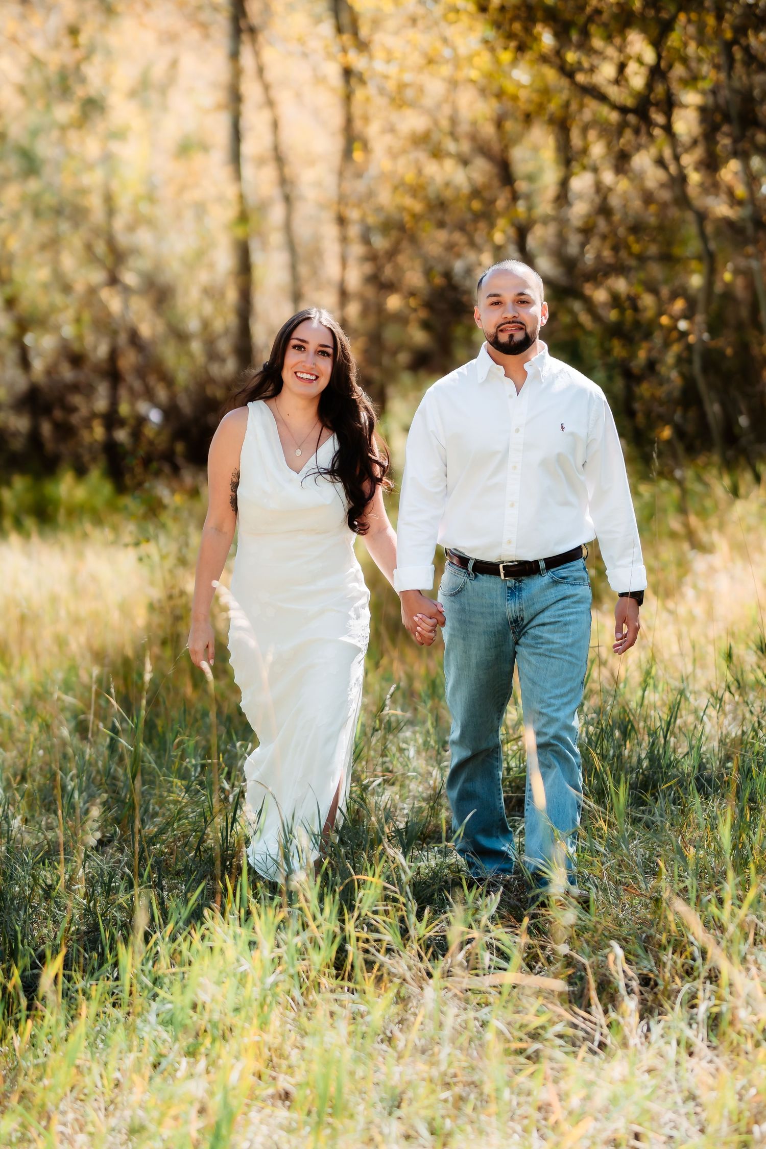 A couple walks through tall grass in a sun-drenched meadow wearing white and blue casual attire.