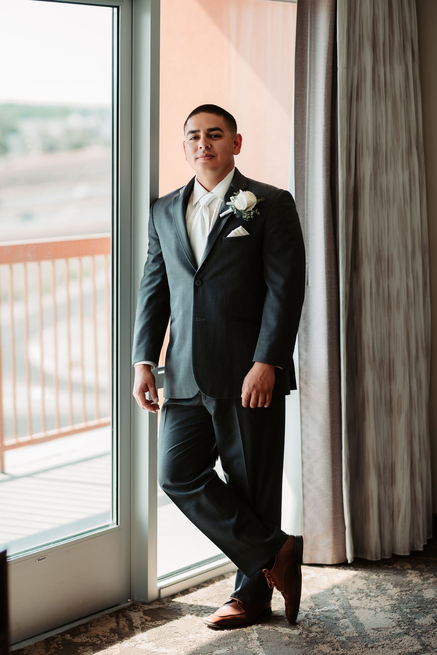 A groom in a classic black suit and white shirt stands by large windows in a formal wedding venue.