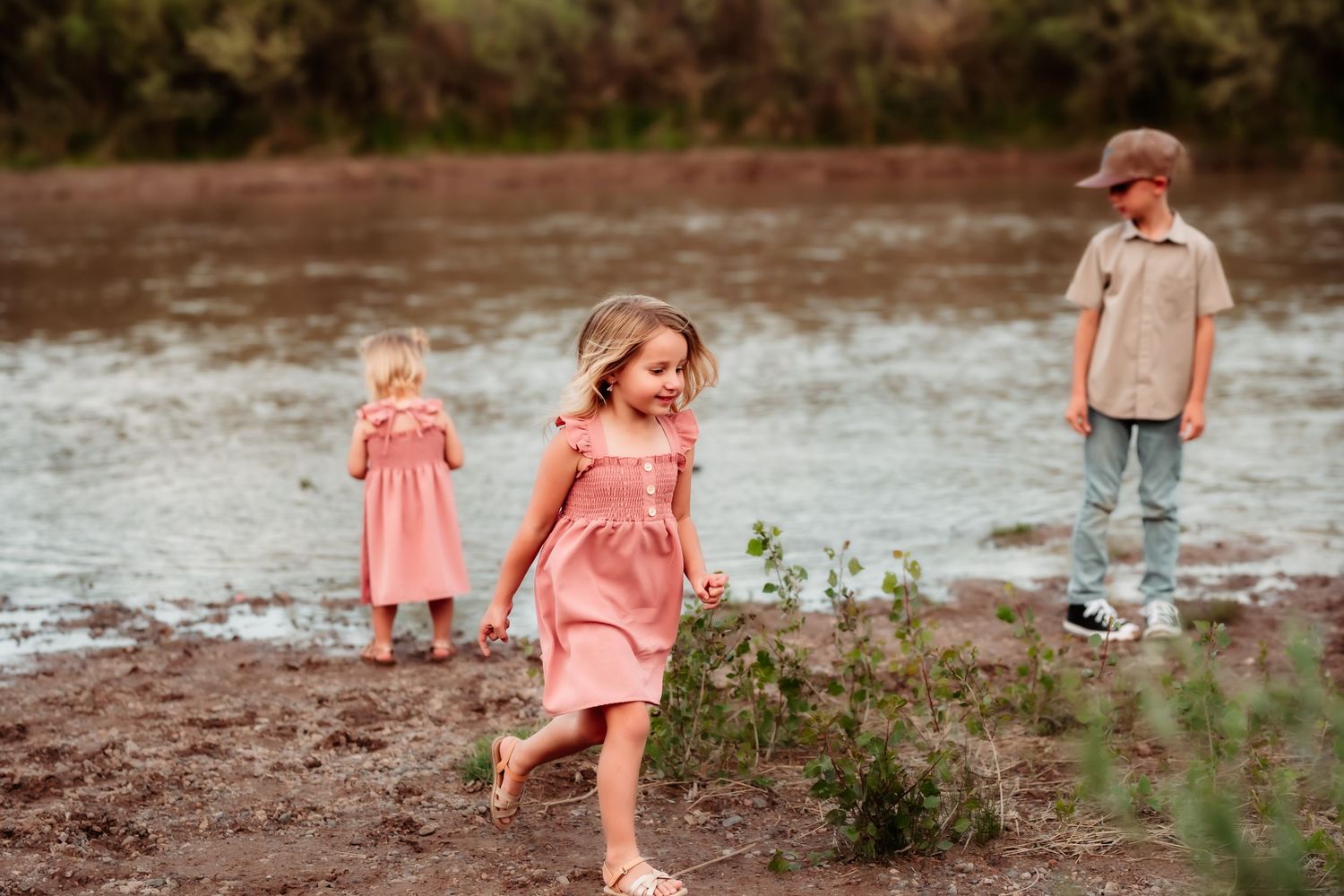 Children playing by a muddy riverbank wearing pink dresses and casual wear on an autumn afternoon.
