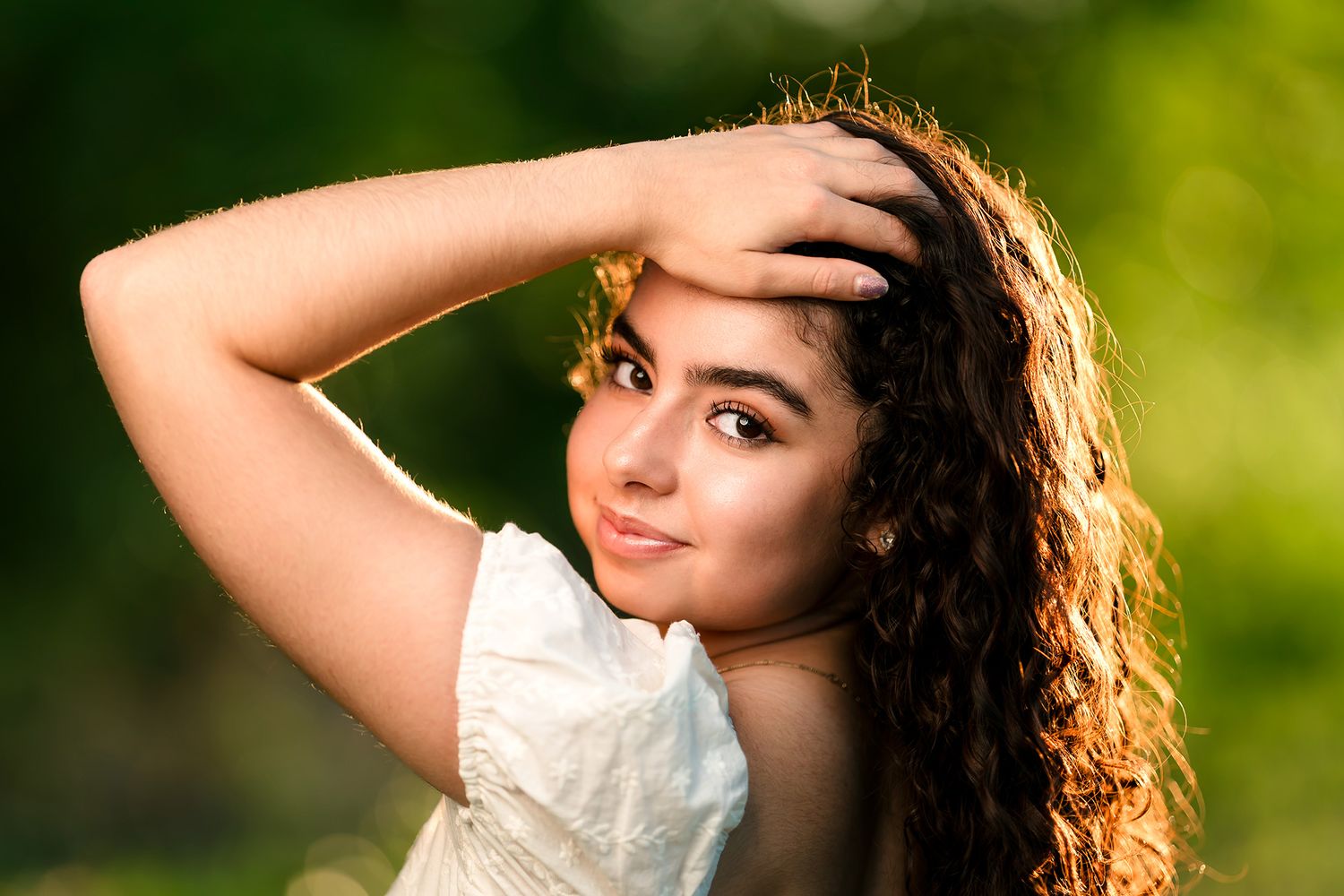 Young woman with curly hair poses outdoors in white dress, hand on head, with lush green bokeh background.