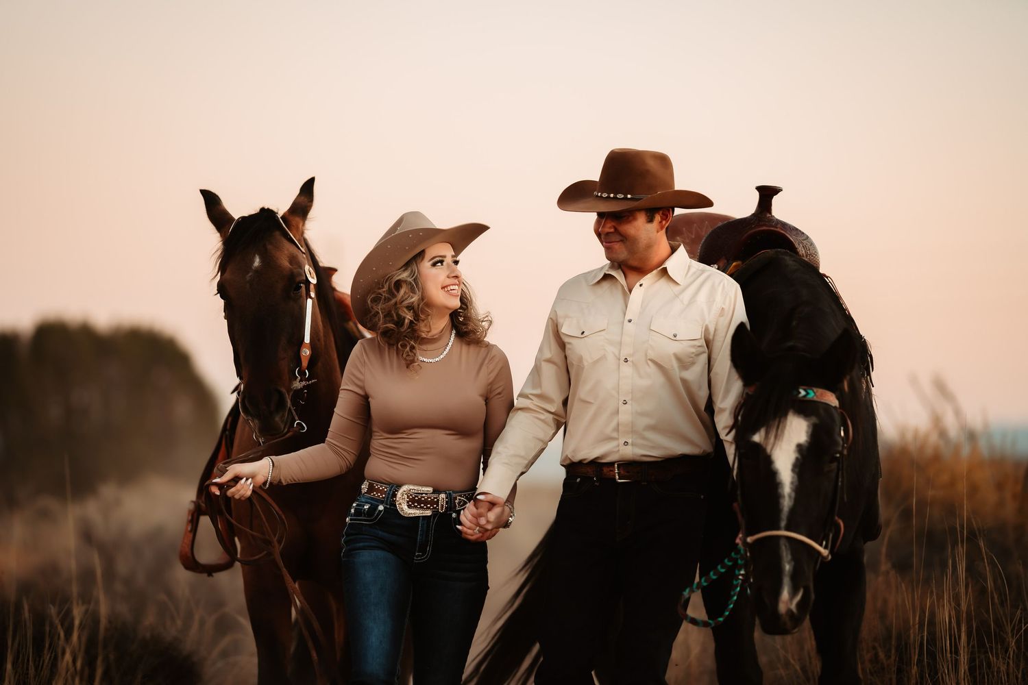 A couple poses with their horses in a rustic field during golden hour sunset.