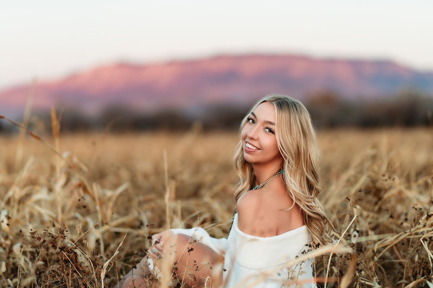 Smiling blonde woman sitting in golden wheat field with purple mountain sunset backdrop.