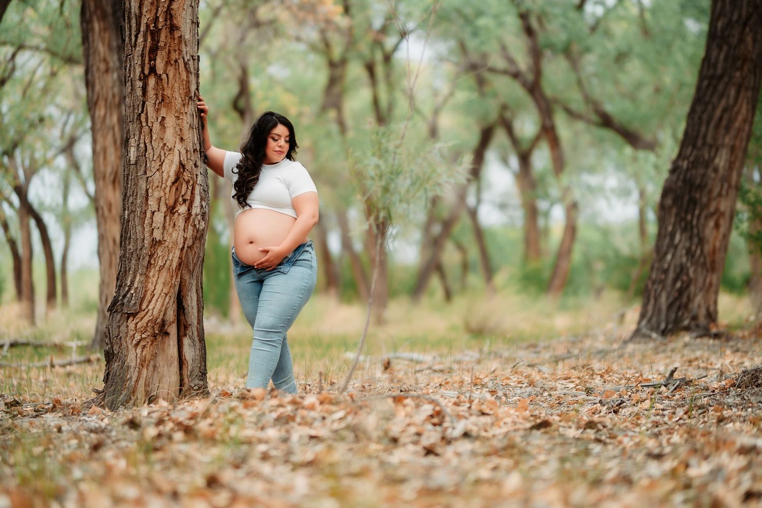 An expectant mother in jeans stands by a tree trunk in a serene autumn forest setting.