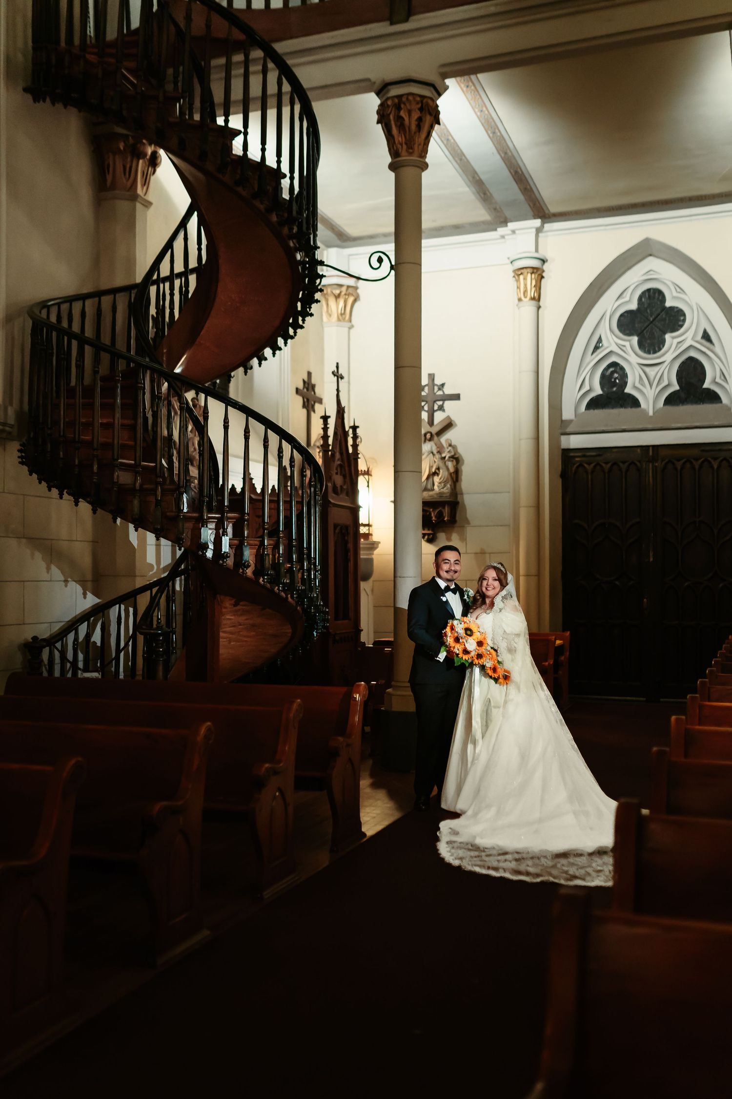 A couple poses on an elegant wooden spiral staircase with Gothic architectural details in a historic interior setting.