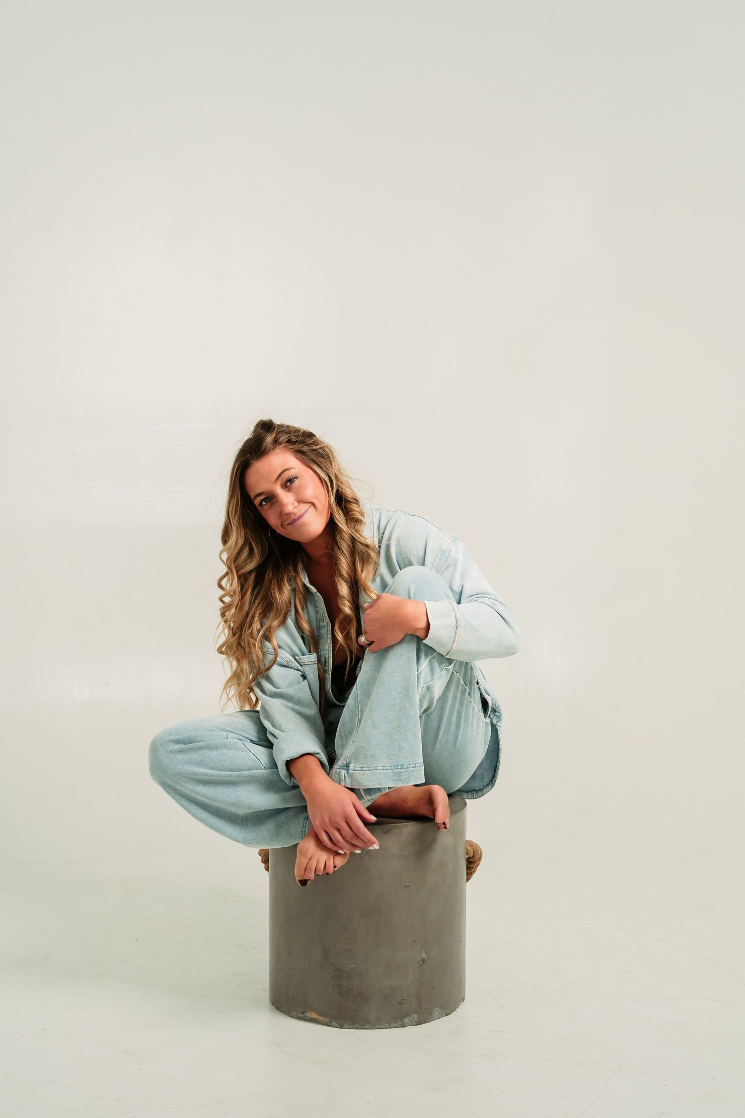 A woman in light blue denim poses expressively around a gray concrete cylinder in a studio.