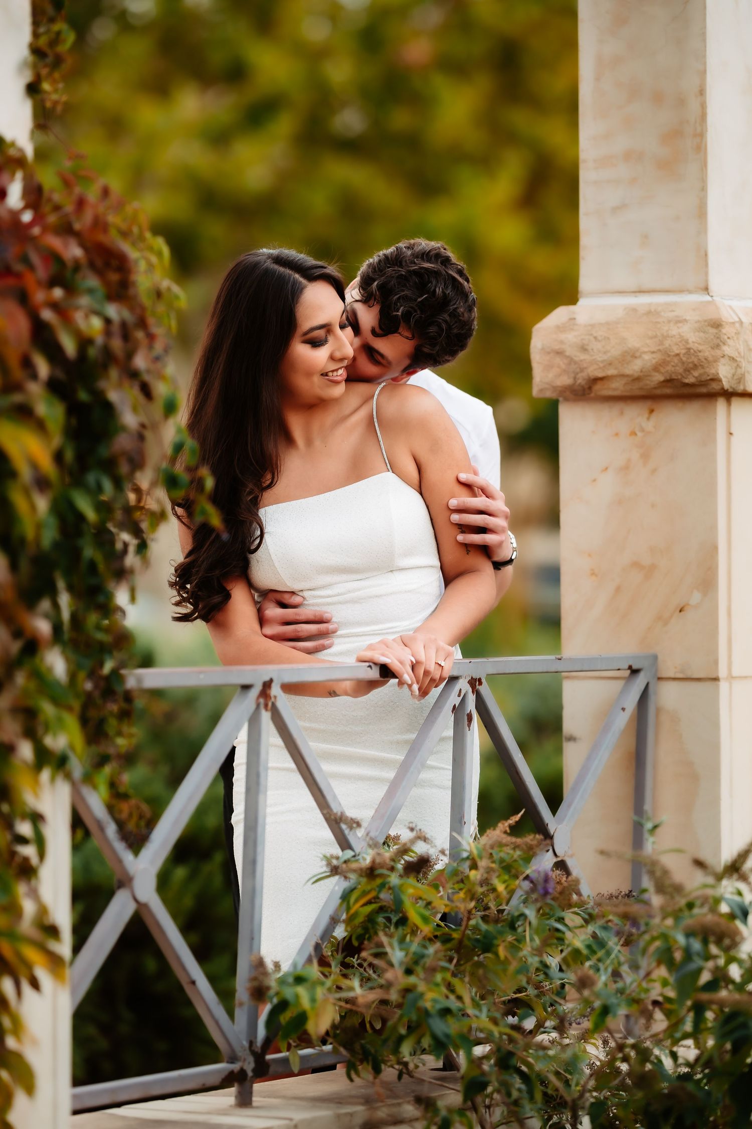 Romantic couple embraces on garden balcony with autumn foliage, bride wearing elegant white strapless wedding dress.