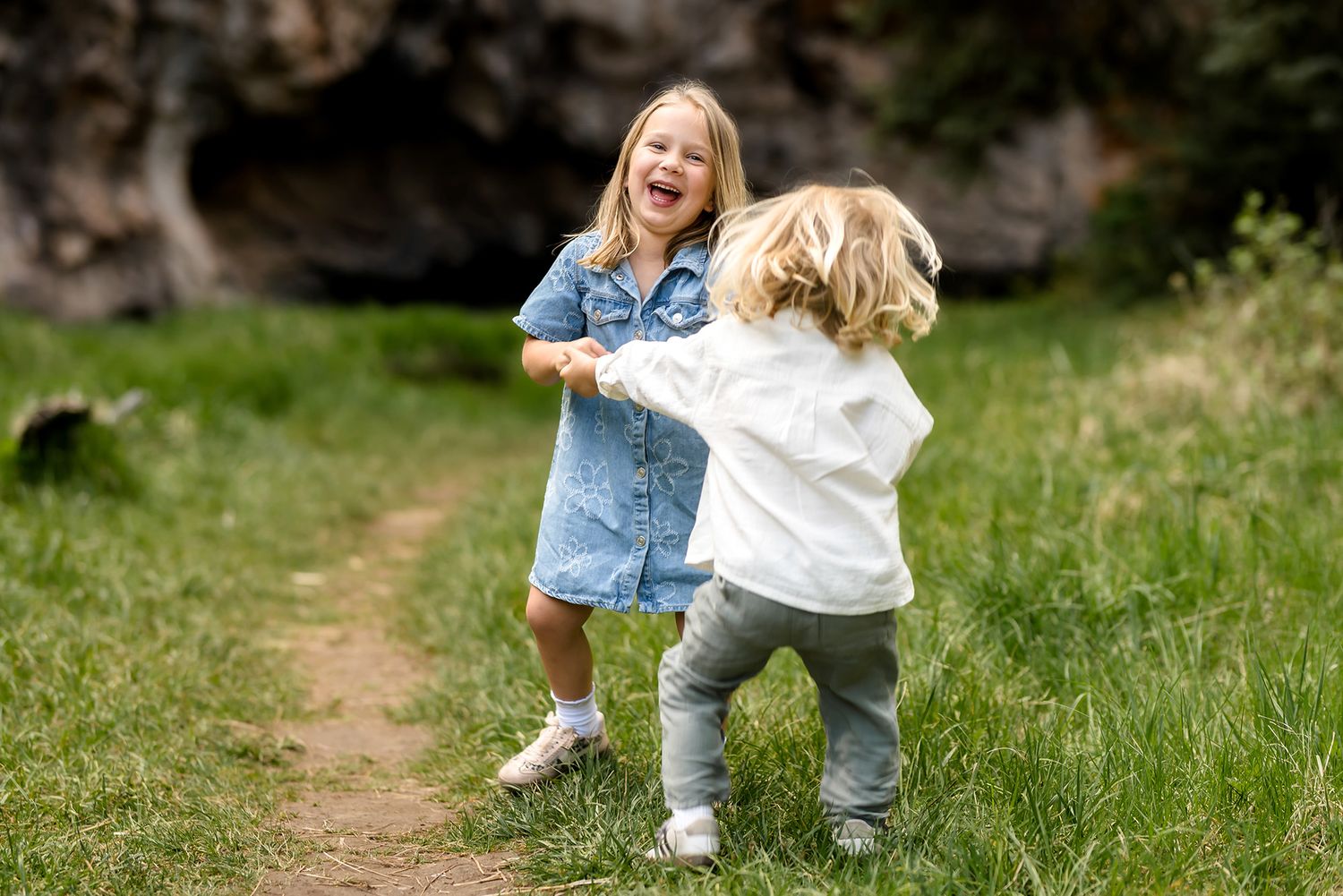 Two young children laughing and playing together on a grassy trail surrounded by nature.