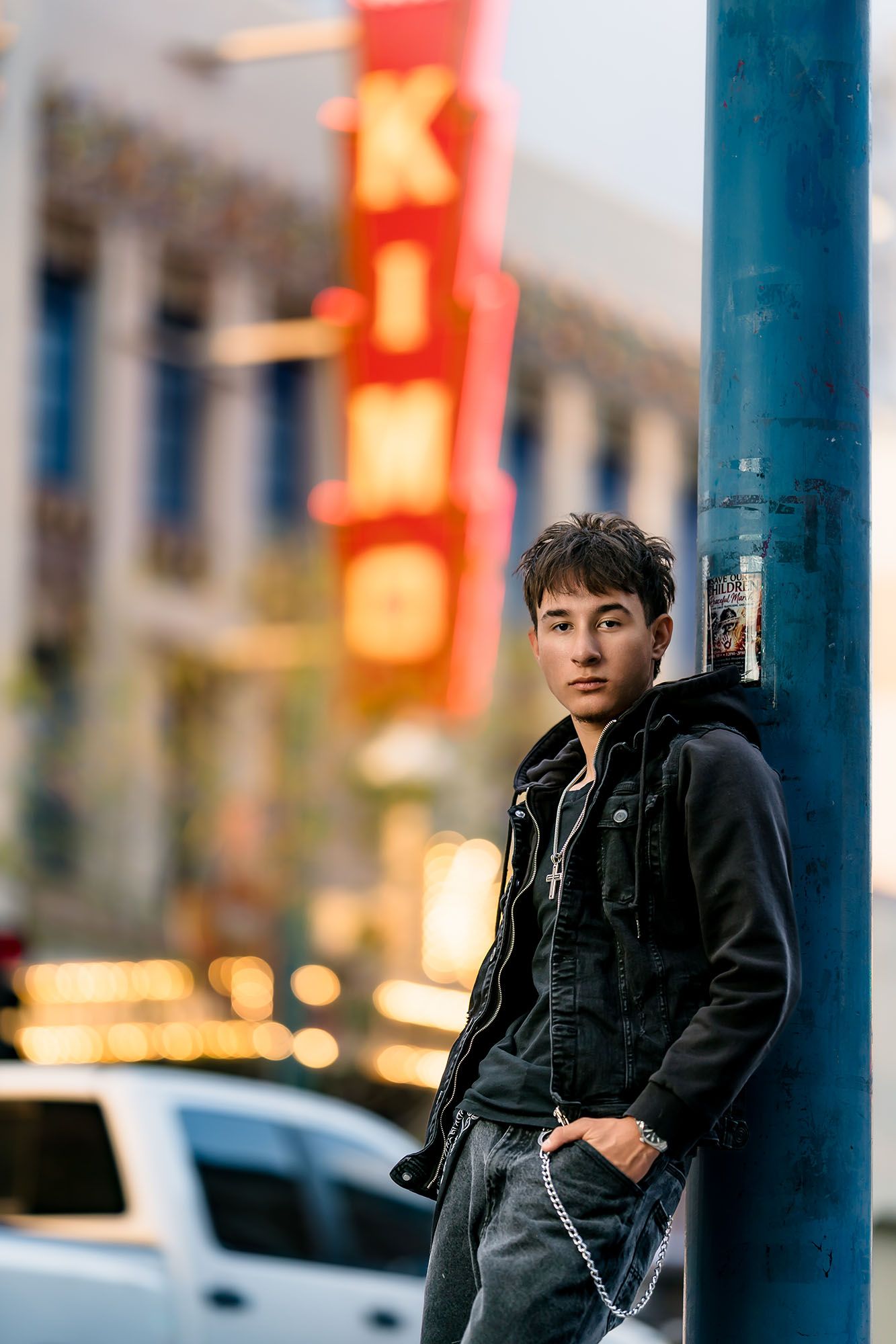 Young Asian man in black jacket leaning against blue pole on urban street with King theater neon sign in background.