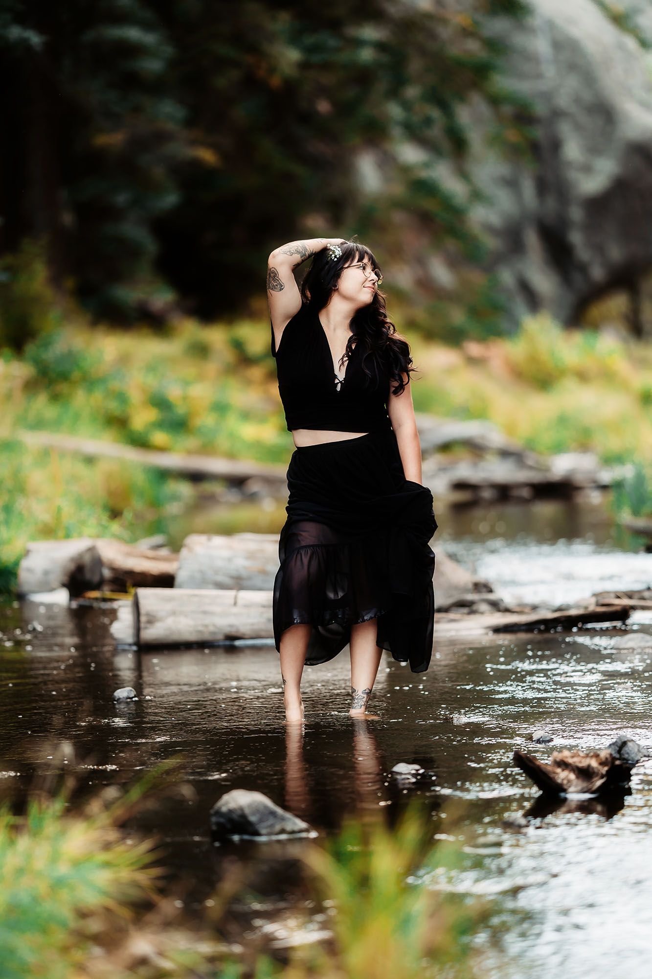 Woman in black outfit stands barefoot in a shallow stream surrounded by nature.