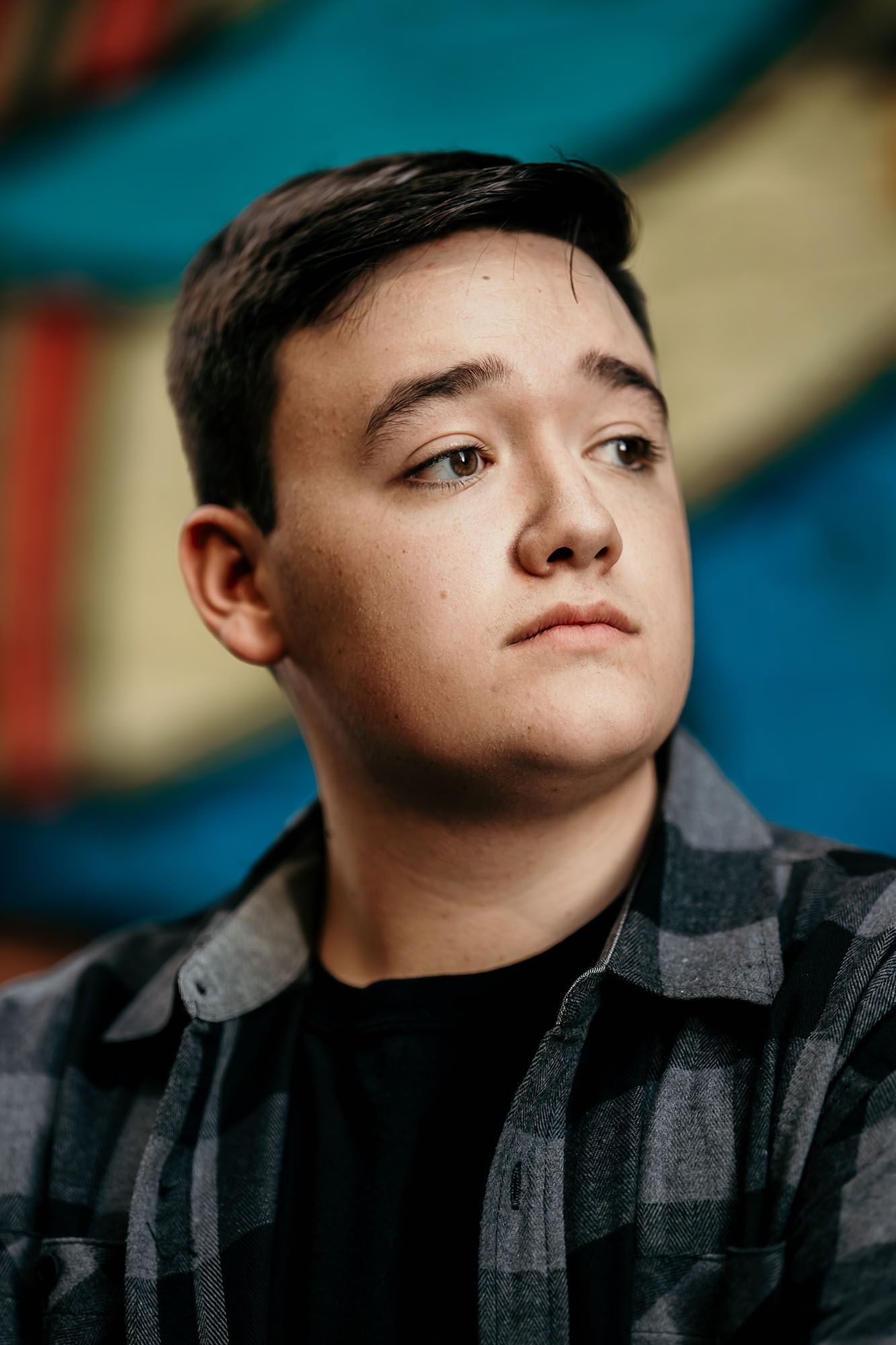 A young man in a plaid shirt poses thoughtfully against a colorful graffiti wall background.