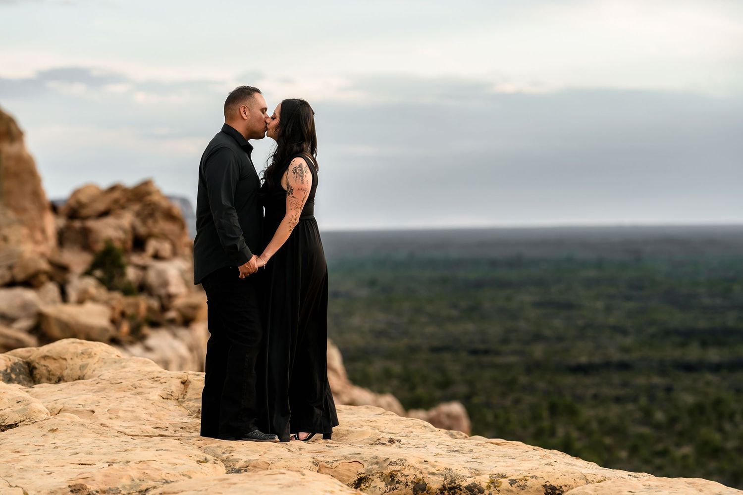 Couple in matching black outfits sharing a kiss on rocky overlook with vast green landscape behind them.