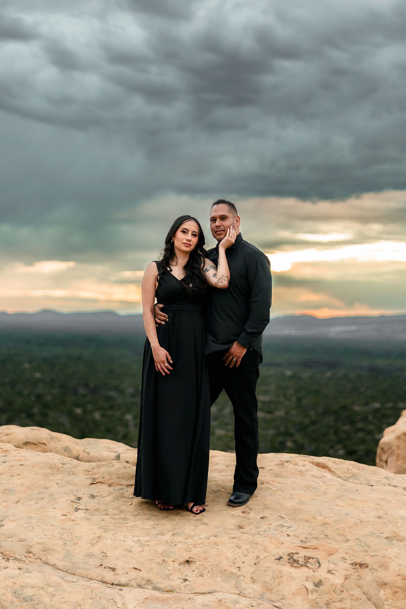 Couple in matching black outfits posing on rocky cliff at sunset with dramatic stormy sky and landscape.