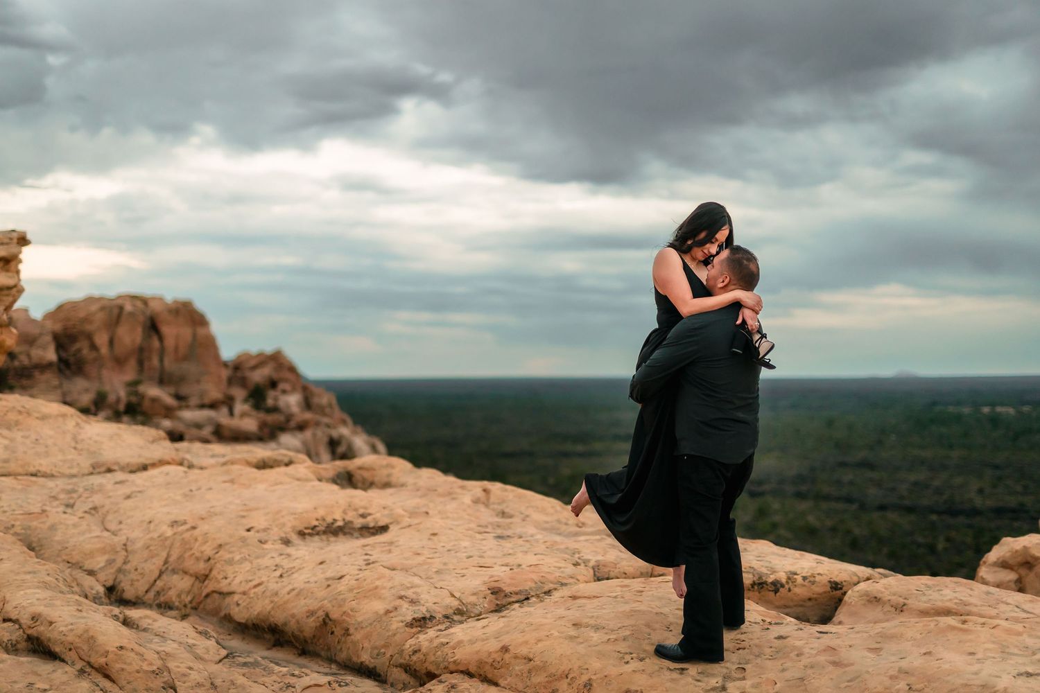 Man lifting woman in black dress on rocky overlook under dramatic cloudy sky with forest below.