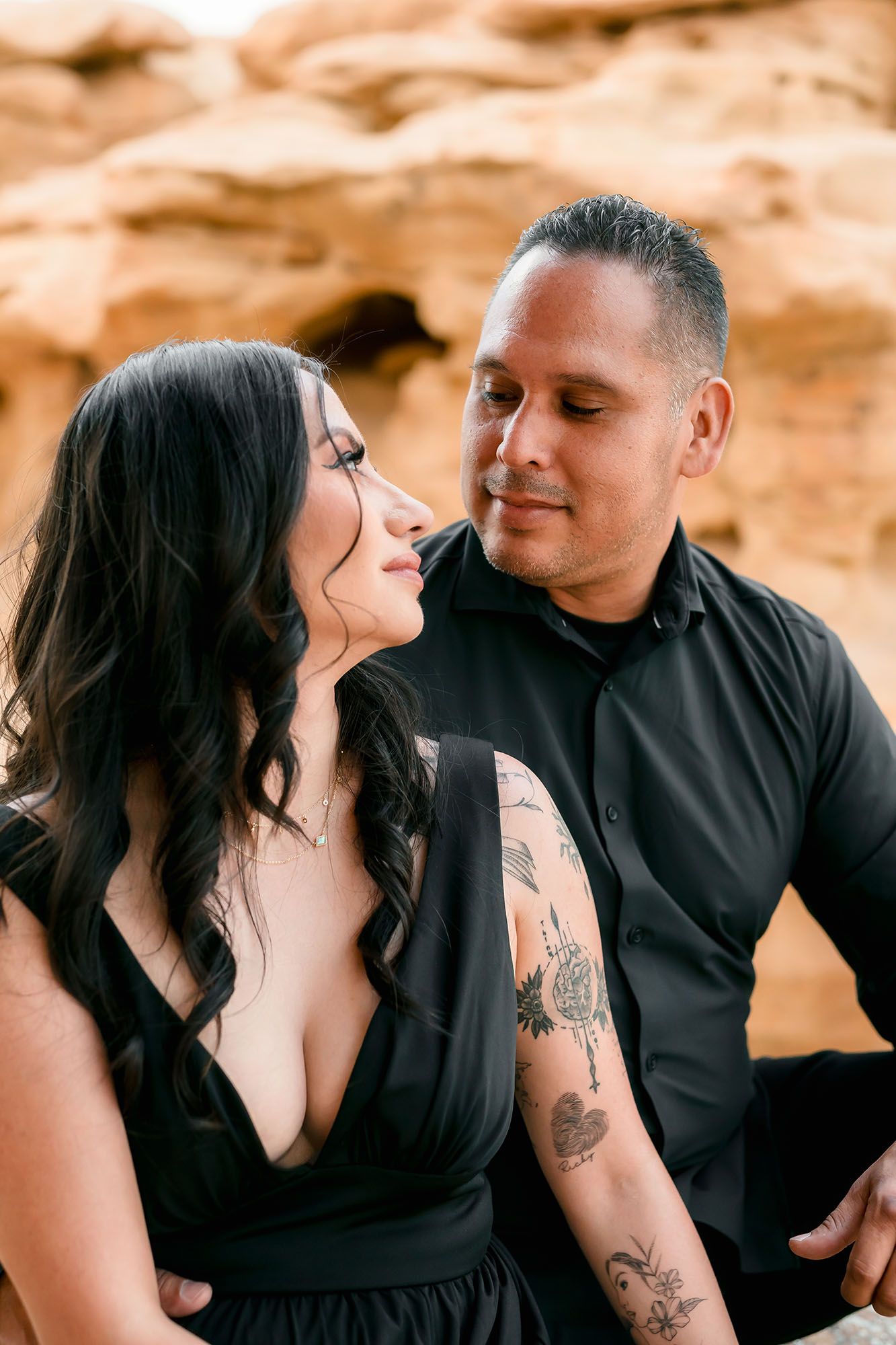 Tattooed couple in black clothing sharing an intimate close-up moment against sandy rock backdrop.