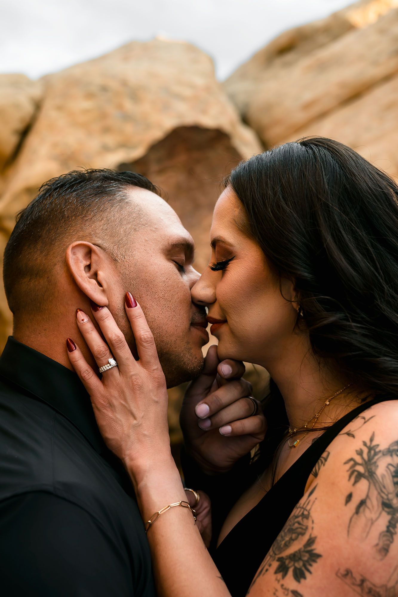 Engaged couple kissing near desert rocks, woman with tattoo and red nails holding mans face tenderly.